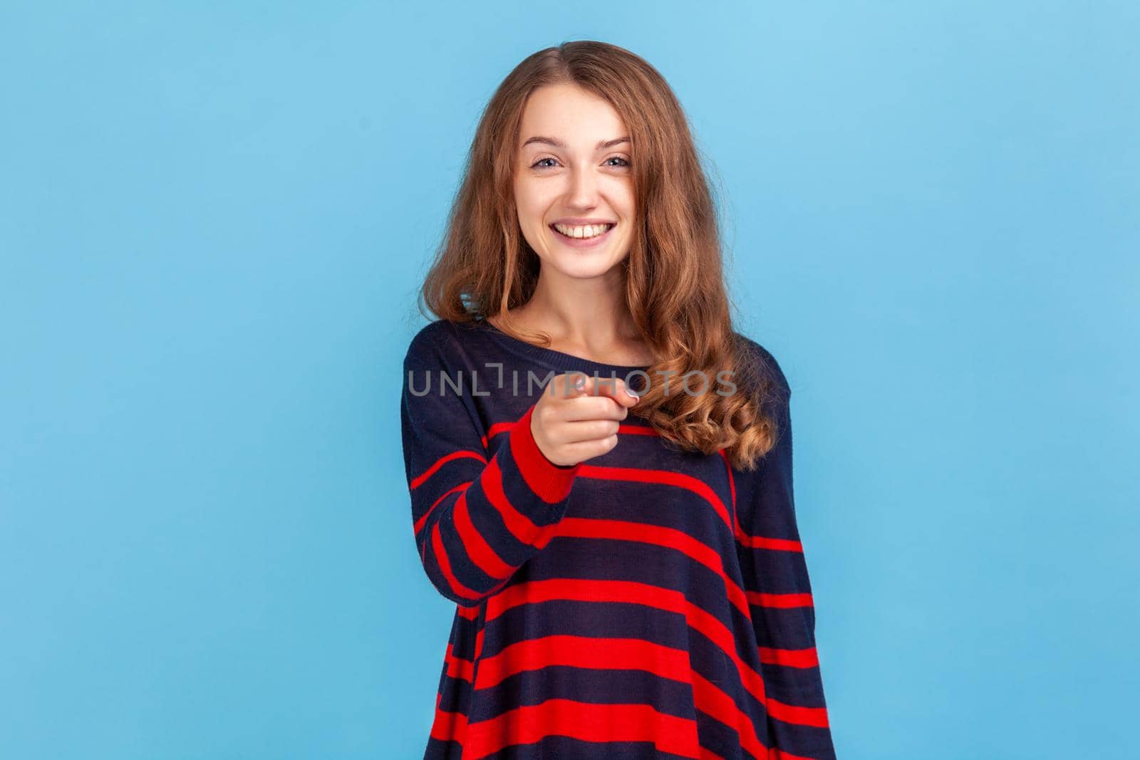 Hey you. Happy woman wearing striped casual style sweater pointing to camera and looking with charming toothy smile, indicating finger, making choice. Indoor studio shot isolated on blue background.