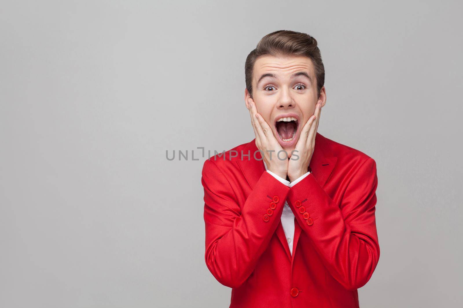 Oh my god. Portrait of extremely amazed gentleman with stylish hairdo in red tuxedo and bow tie holding hands on cheeks and looking at camera with shocked eyes. indoor studio shot, gray background
