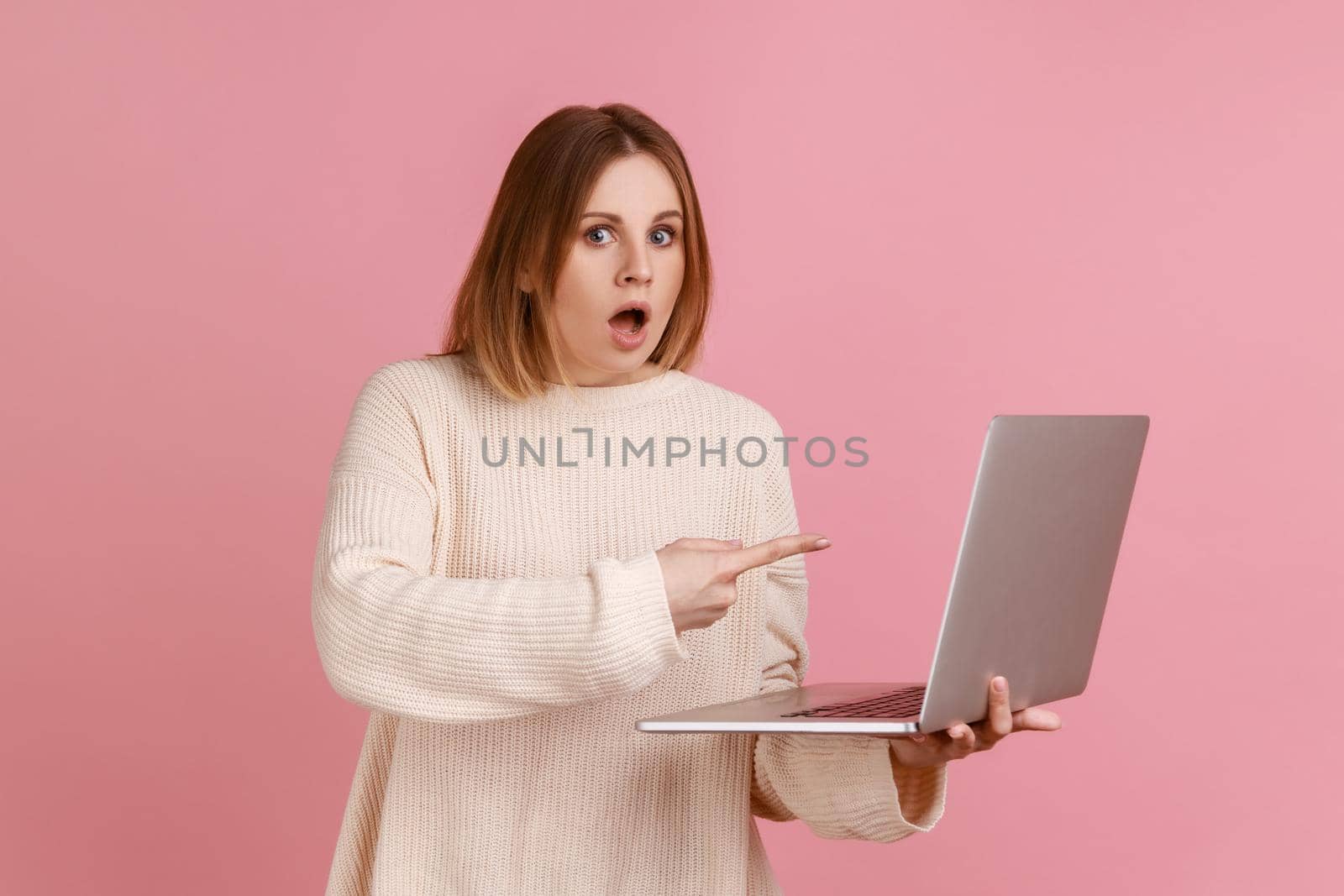 Shocked young adult blond woman working on laptop compute, looking at camera and pointing at display by Khosro1