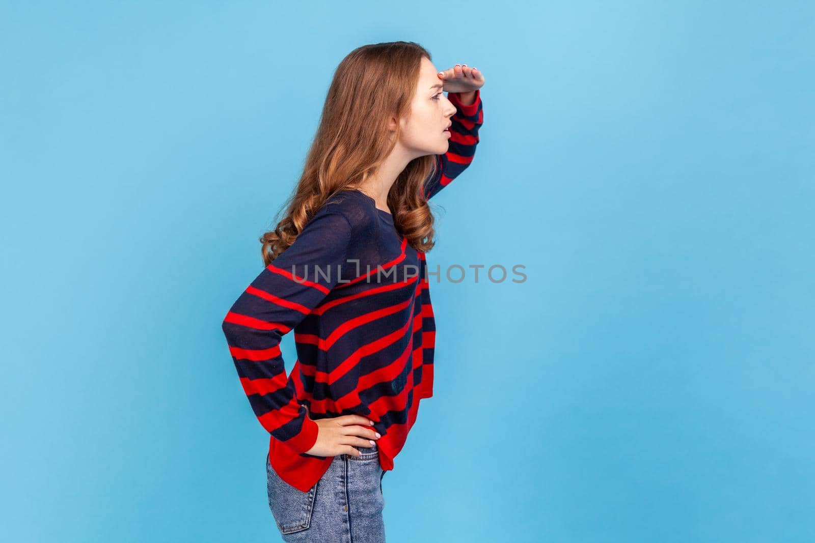 Side view portrait of woman wearing striped casual style sweater keeps hand near forehead looks far away searches something on horizon. Indoor studio shot isolated on blue background.