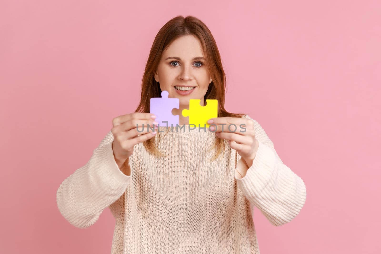 Portrait of smiling positive attractive blond woman holding yellow and purple puzzle pieces, solving tasks, looking at camera, wearing white sweater. Indoor studio shot isolated on pink background.