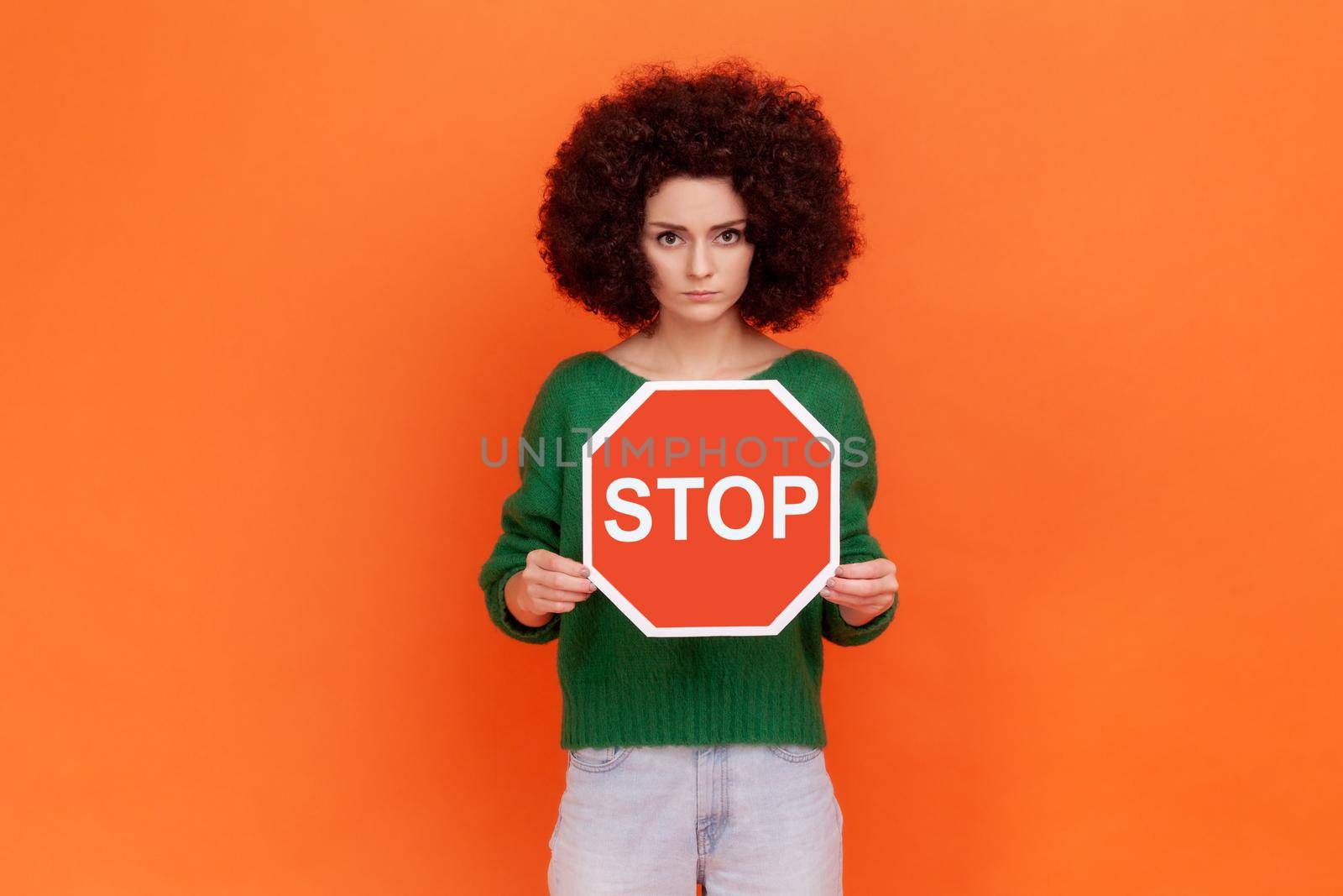 Serious strict woman with Afro hairstyle wearing green casual style sweater holding red stop sign in hands, looking at camera, forbidden actions. by Khosro1