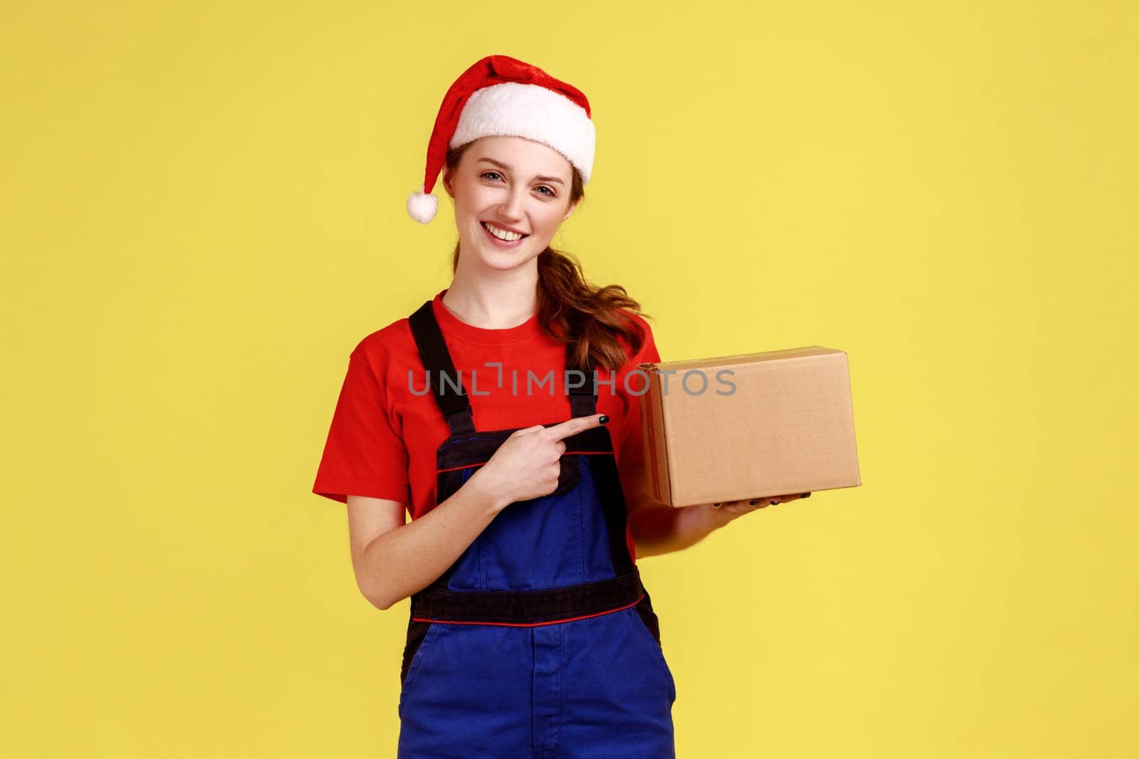 Pleased delivery woman holding pointing at parcel, fast delivering of presents for winter holiday, wearing blue overalls and santa claus hat. Indoor studio shot isolated on yellow background.