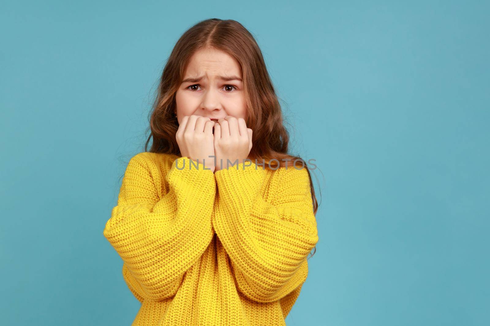 Little girl biting nails with scared big eyes, feeling worried, anxious about childish mistake, wearing yellow casual style sweater. Indoor studio shot isolated on blue background.