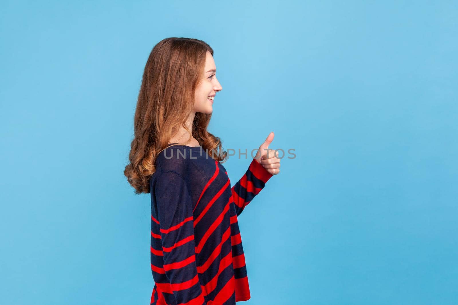 Side view of smiling woman wearing striped casual style sweater looking away, showing thumb up, expressing approval, excellent feedback. Indoor studio shot isolated on blue background.