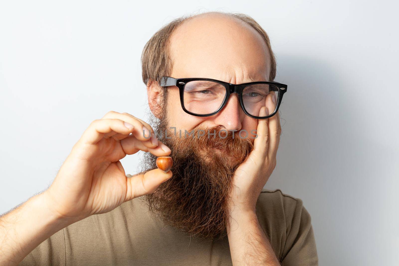 Portrait of unhealthy male suffering terrible toothache after cracking hazel, needs dental treatment, bald bearded man wearing T-shirt and glasses. Indoor studio shot isolated on gray background.