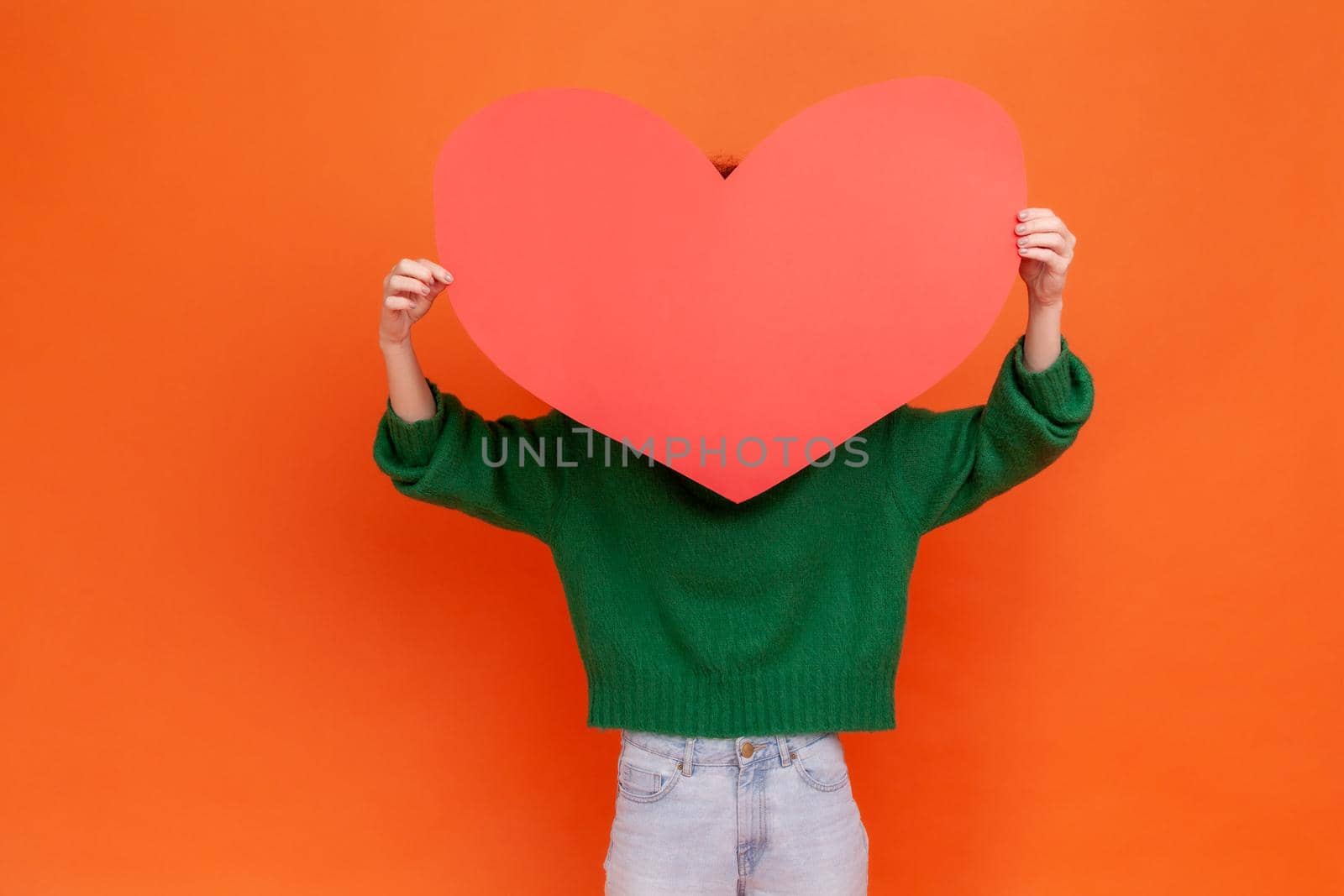 Unknown woman wearing green casual style sweater holding big red paper heart, hiding behind love huge love symbol, shy to express her feelings. Indoor studio shot isolated on orange background.