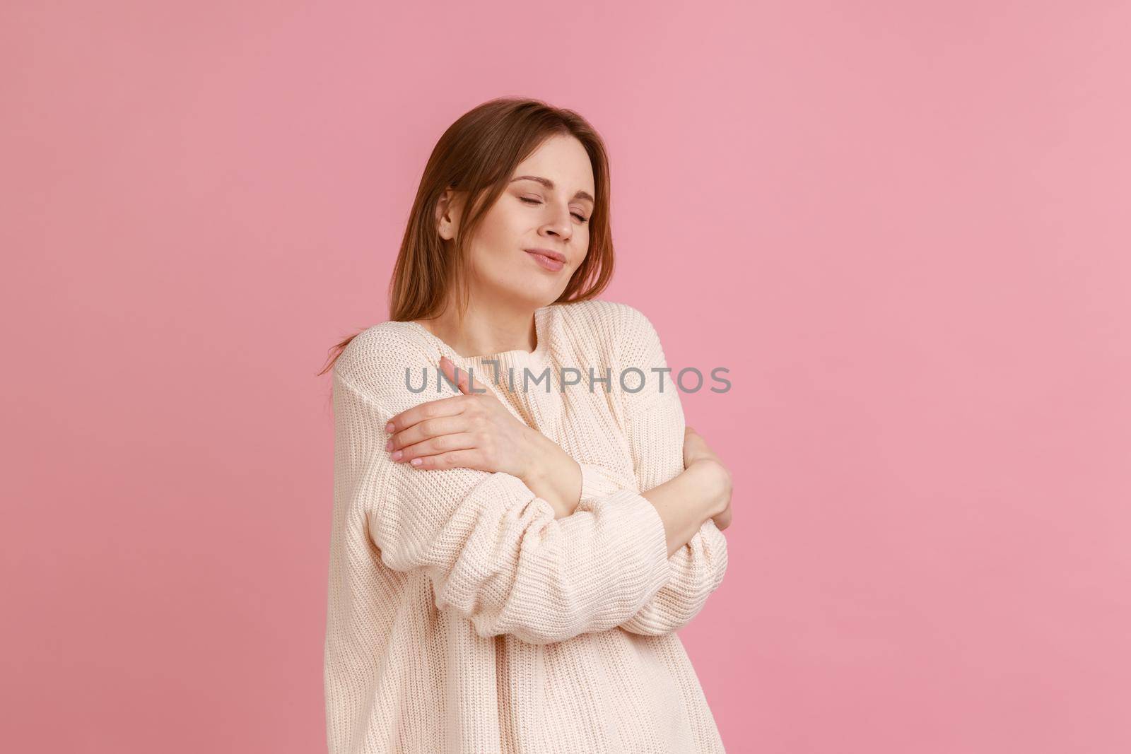 I love myself. Portrait of woman hugging herself and smiling, feeling comfortable and fulfilled, narcissistic egoistic person, wearing white sweater. Indoor studio shot isolated on pink background.