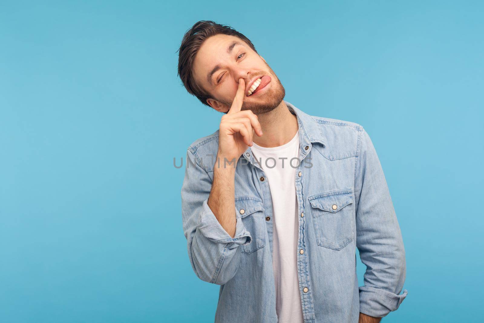 Portrait of funny man wearing denim shirt, putting finger into his nose and showing tongue, fooling around, bad habits, disrespectful behavior. Indoor studio shot isolated on blue background.