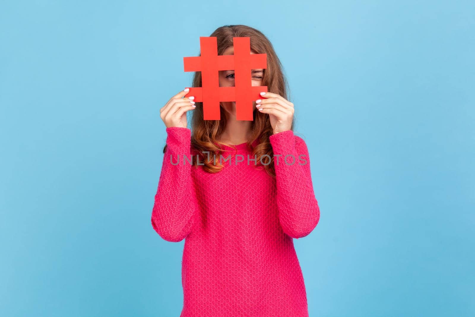 Portrait of attractive young adult woman wearing pink pullover, looking through big red hashtag symbol, popular internet idea, viral content. Indoor studio shot isolated on blue background.