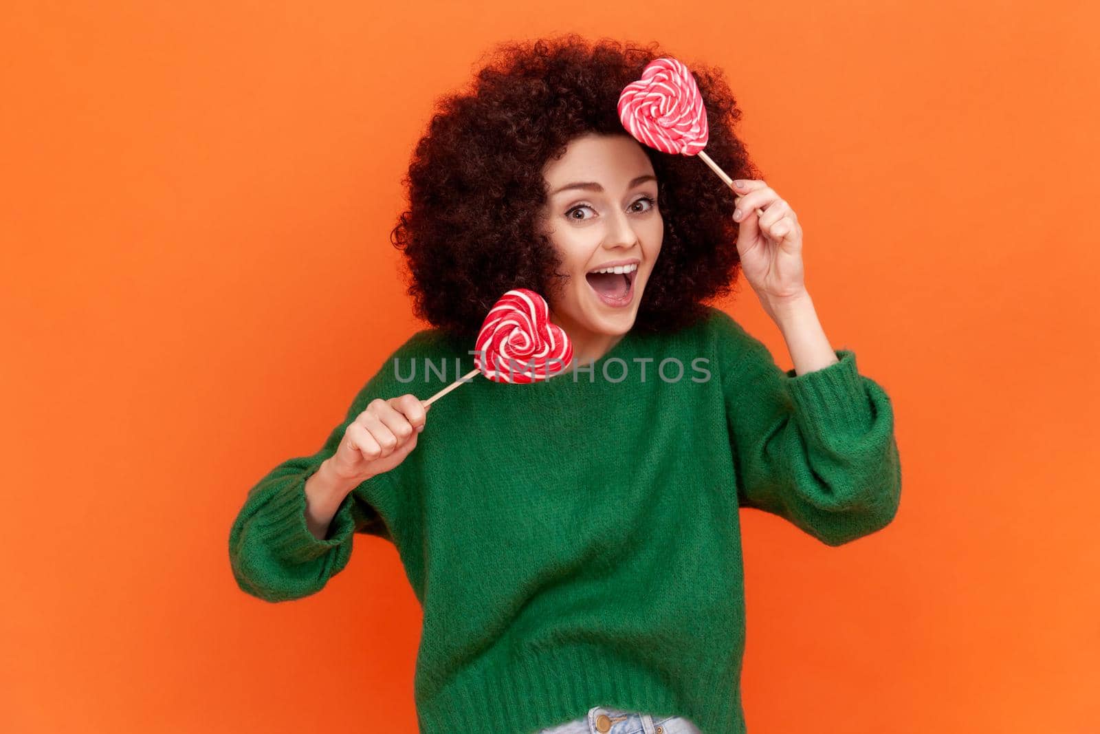 Positive satisfied woman with Afro hairstyle wearing green casual style sweater holding two heart shape lollipops, having fun, funny expressions. by Khosro1