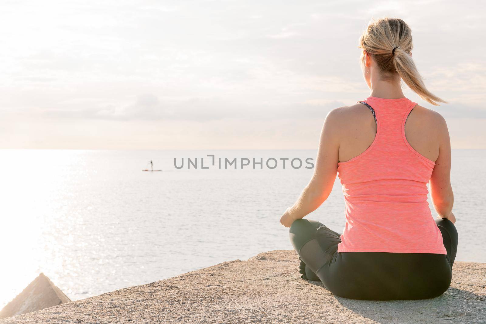 back of an unrecognizable blonde woman practicing yoga at sunrise sitting in the lotus position in front of the sea, concept of mental health and relaxation, copy space for text