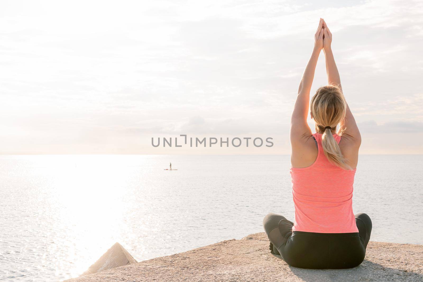 back view of an unrecognizable blonde woman practicing yoga at sunrise sitting in front of the sea, concept of mental health and relaxation, copy space for text
