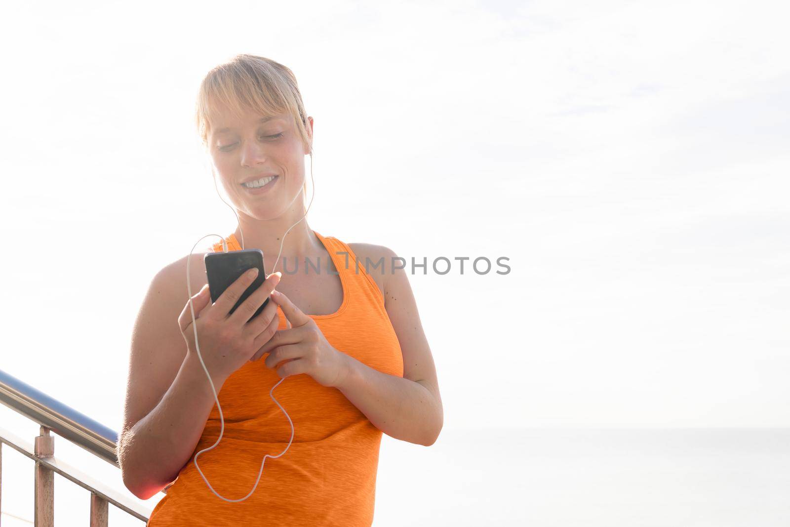 young sporty caucasian girl with earphones listening to music from her mobile phone, technology and healthy lifestyle concept, text copy space and lens flare effect