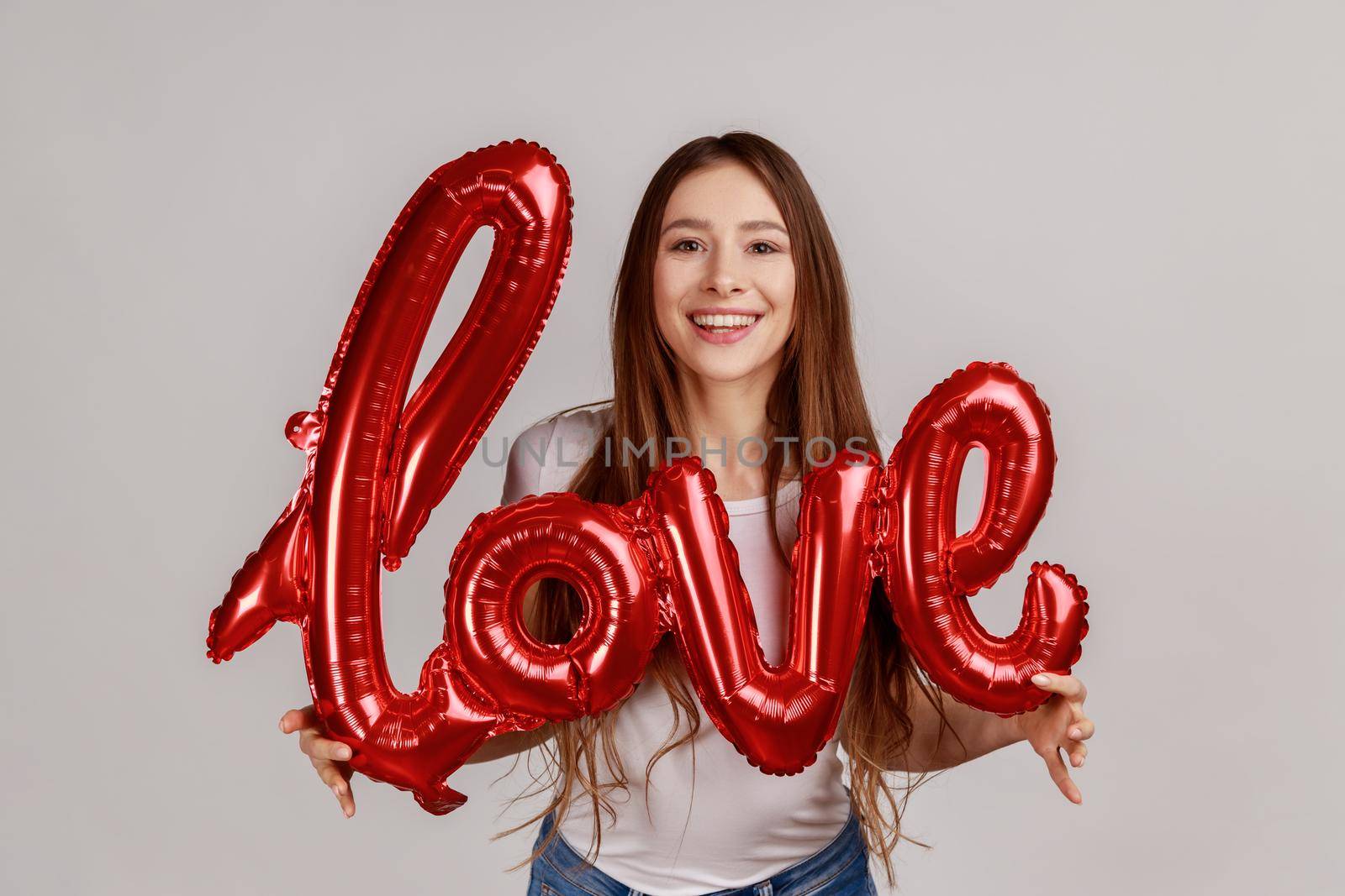 Portrait of beautiful satisfied romantic woman holding foil balloon letters, looking at camera, expressing love, wearing white T-shirt. Indoor studio shot isolated on gray background.