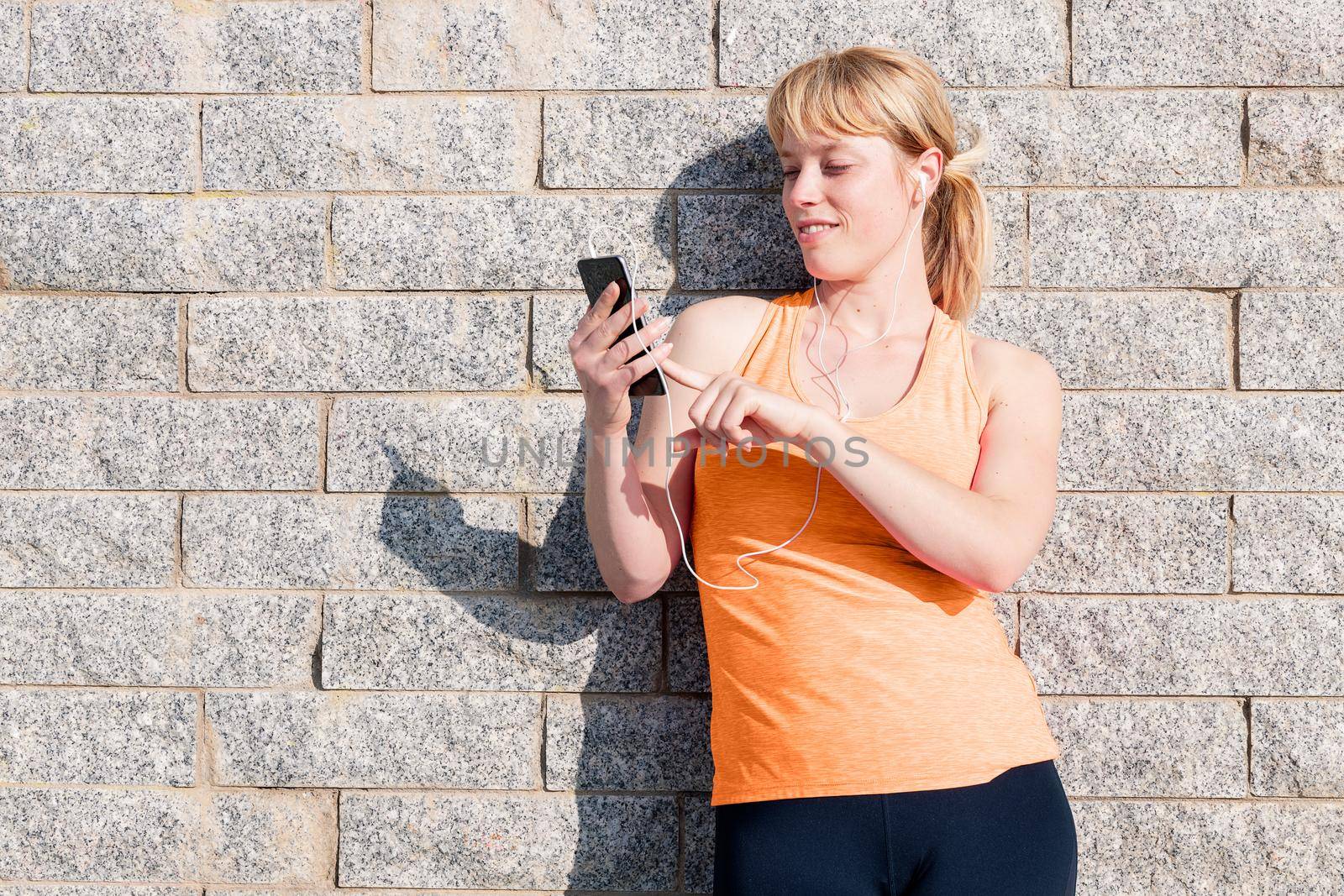 young sporty blonde girl with earphones consulting her mobile phone leaning against a stone wall, technology concept and healthy lifestyle, copy space for text