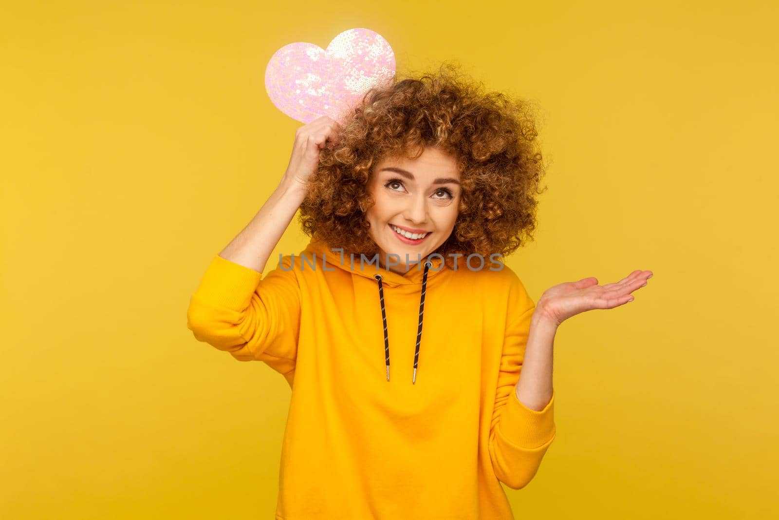 Positive woman with Afro hairstyle and charming smile holding pink paper heart over head and spreading palm aside, wearing casual style hoodie. Indoor studio shot isolated on yellow background.