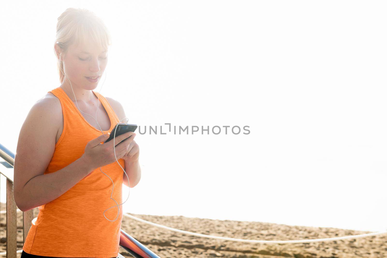 young sporty blonde woman with earphones using her mobile phone by the beach, technology concept and healthy lifestyle, copy space for text and lens flare effect