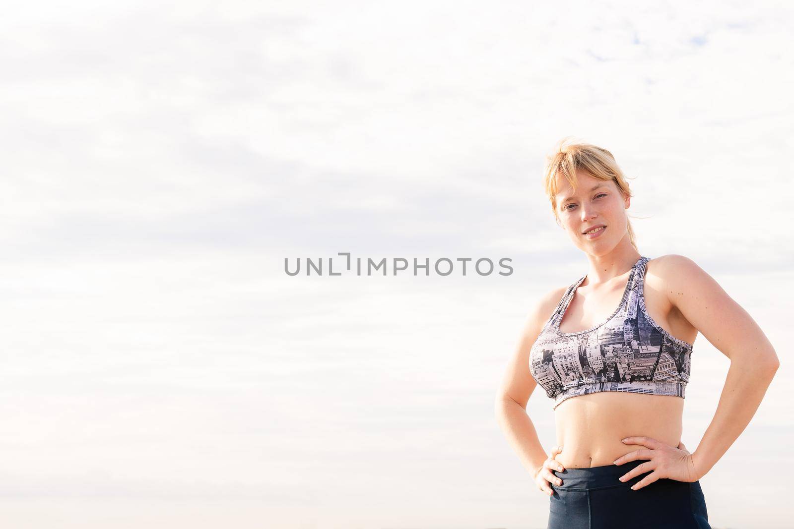 beautiful blonde sportswoman posing powerfully with her hands on her hips with the sky in the background, healthy lifestyle and sports concept, copy space for text