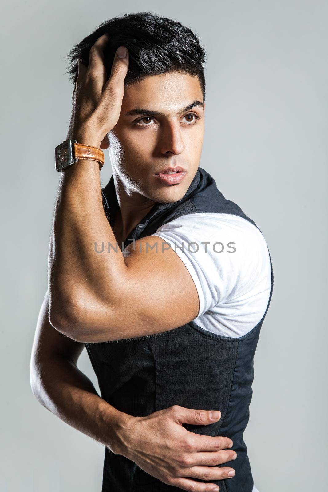 Portrait of fashionable attractive man wearing white T-shirt and black jacket, keeping hand on head, looking away with confident expression. Indoor studio shot isolated on gray background.