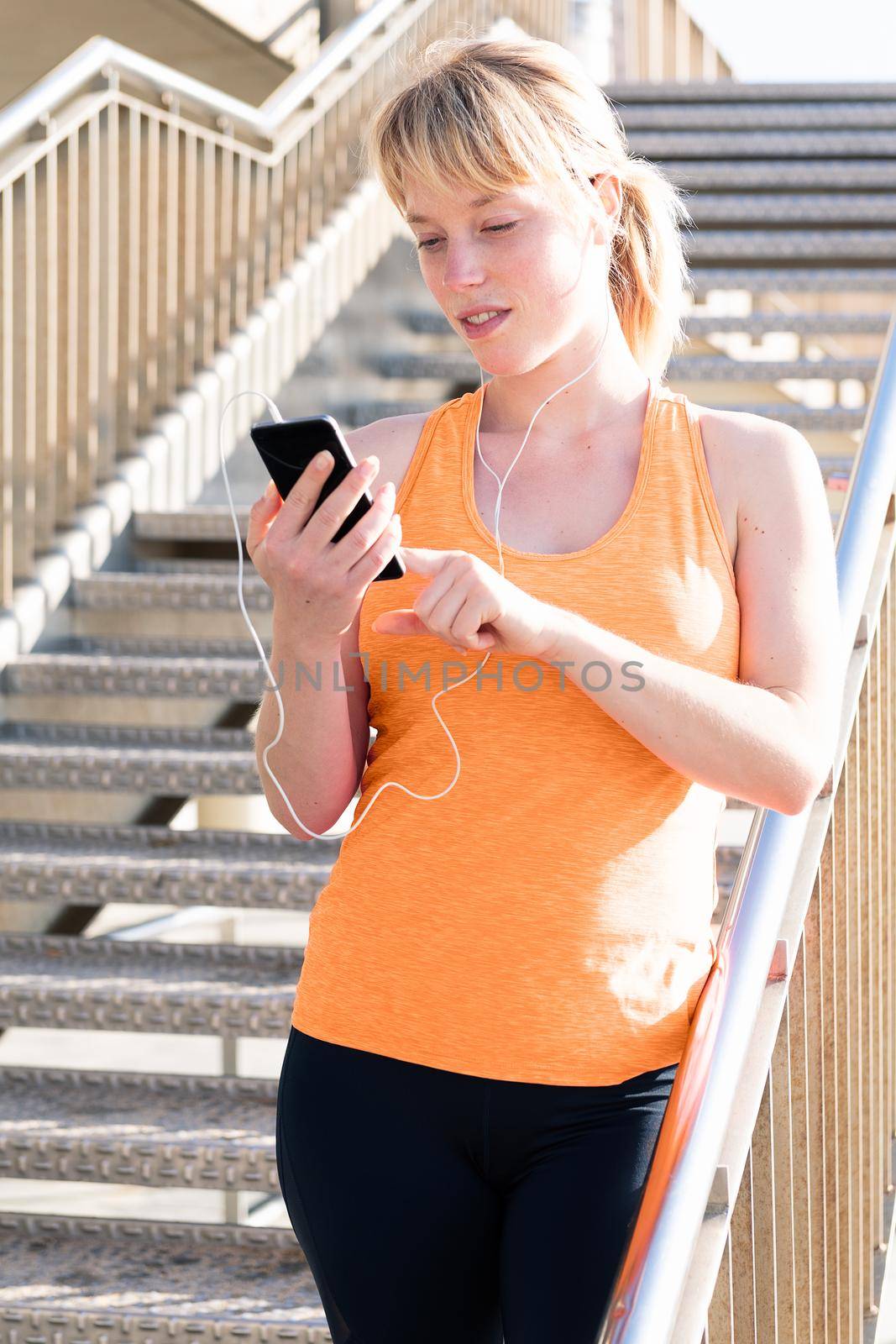vertical photo of a young blonde woman with earphones using her mobile phone next to a staircase, concept of technology and healthy lifestyle