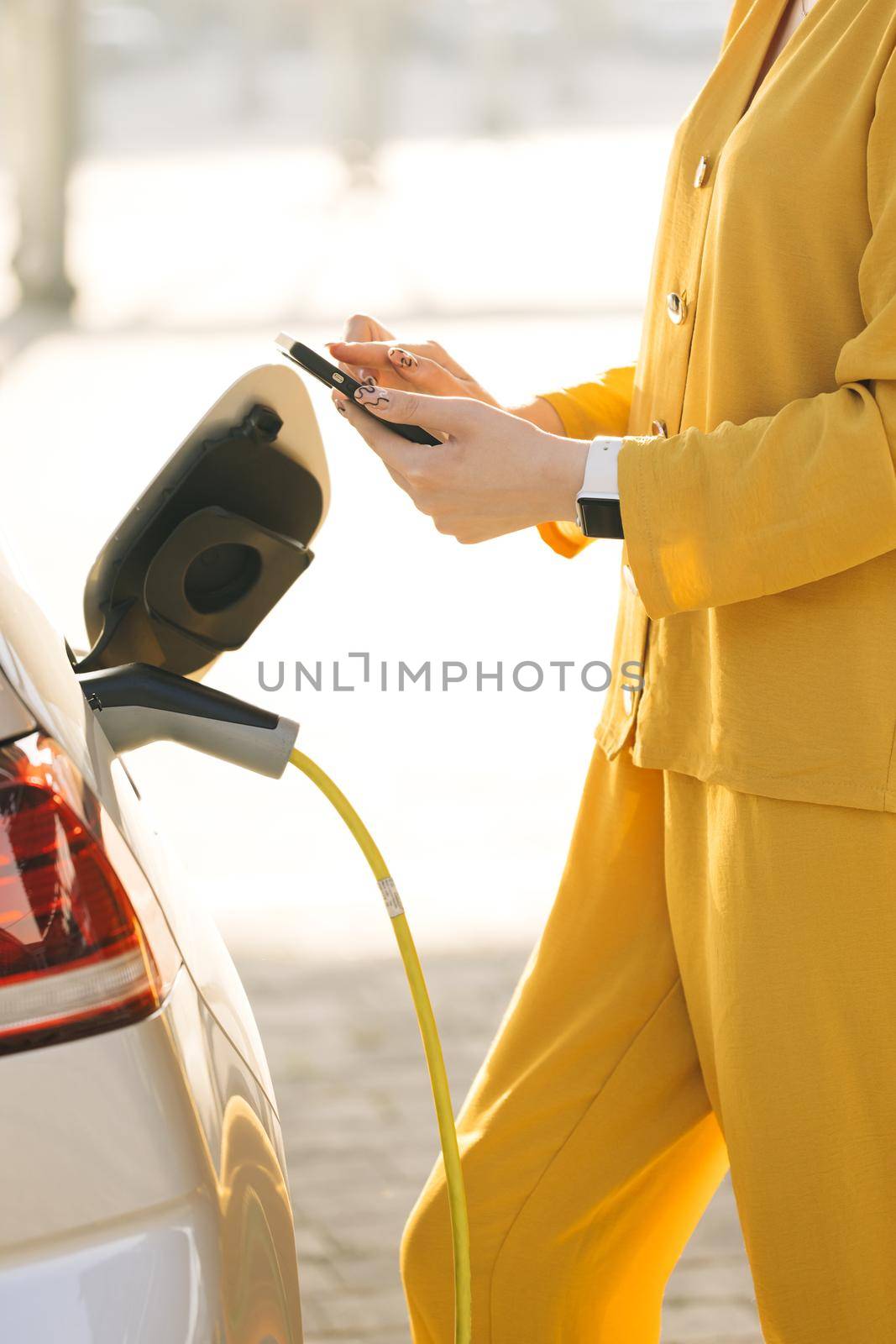 Woman connects an electric car to the charger and adjusts the process of charging the car battery using a cell phone smart phone. Girl plugs power cable to charge electric car in parking lot by uflypro