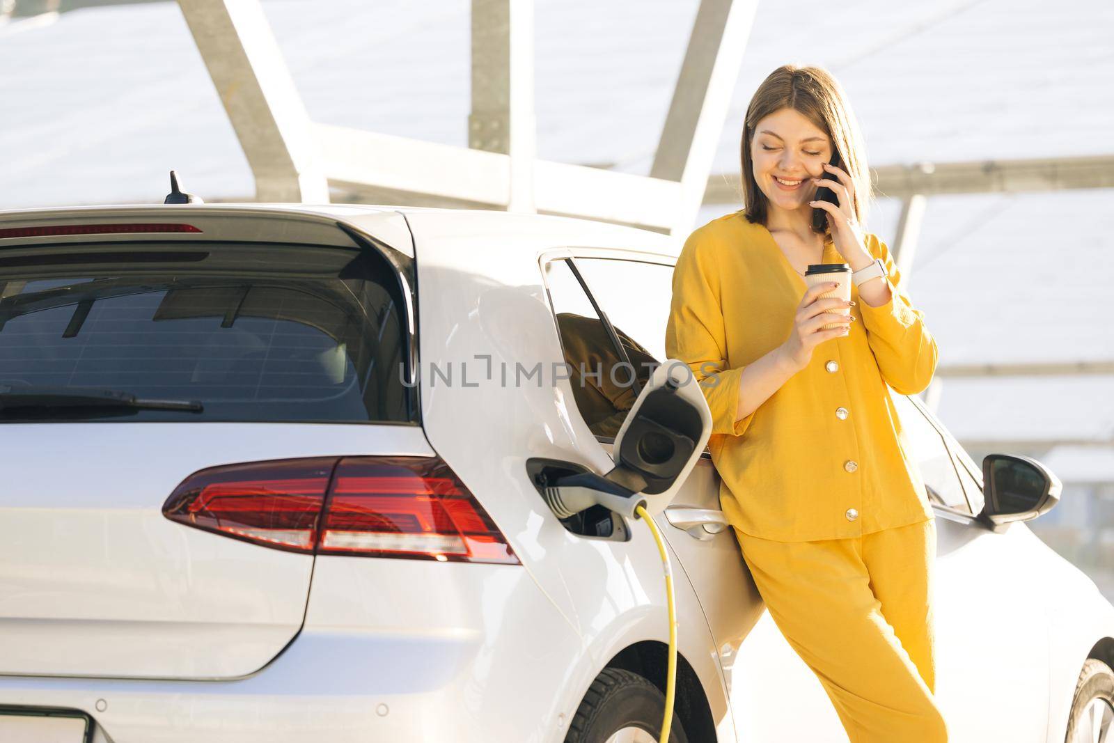 Attractive business woman taking phone call standing near her electric car. Positive woman have talking conversation by phone near her electric car and waits when the vehicle will be charged.