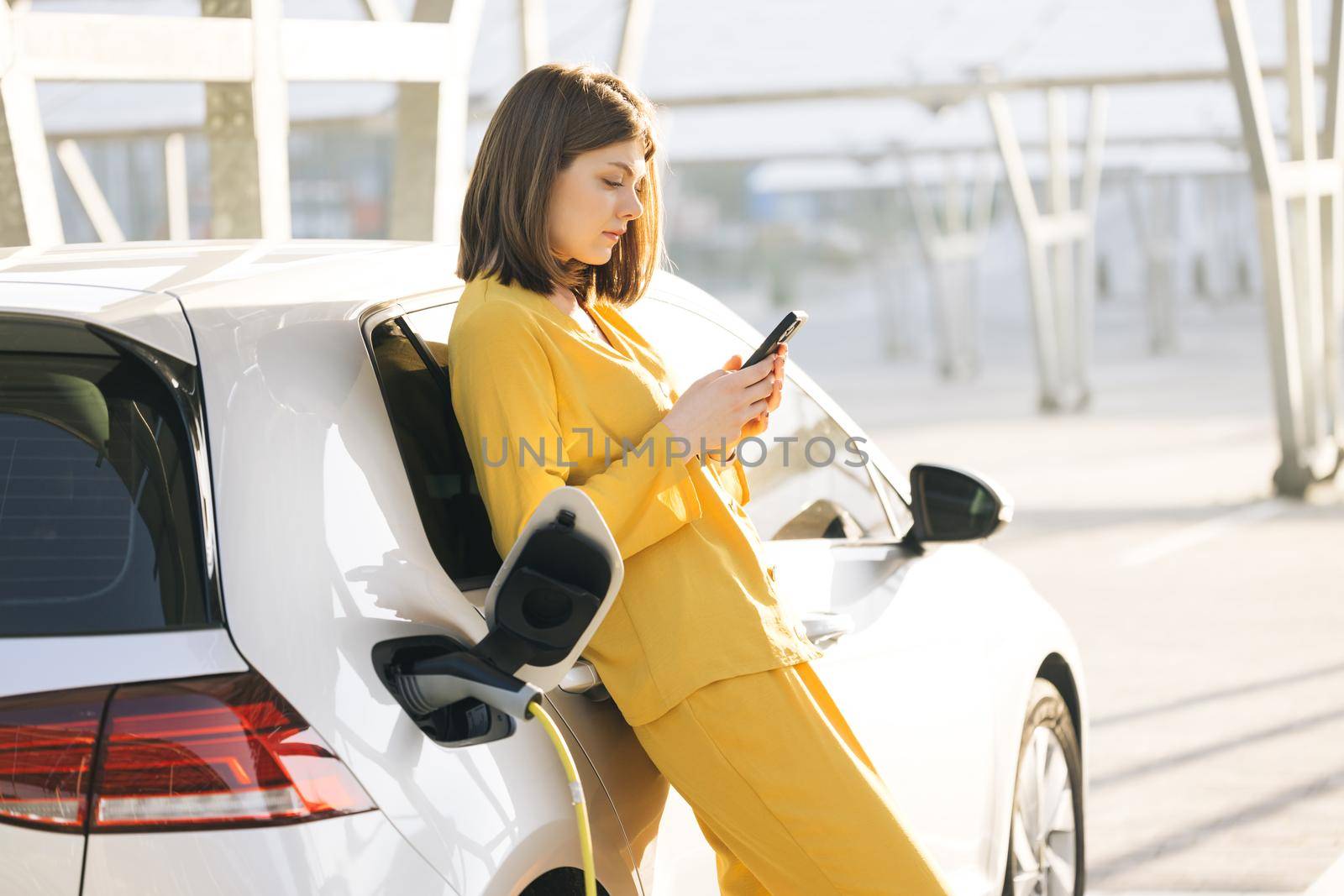 Caucasian woman stands with mobile phone near her electric car and waits when vehicle will charged. Charger plug of an electric car. Electric vehicle charging near an solar power plant. by uflypro