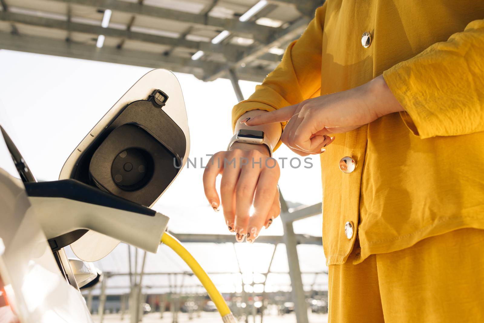 Businesswoman plugging in charging cable to to electric vehicle. Female hand inserts power connector into EV car and charges batteries, uses smartwatch for activates start charging