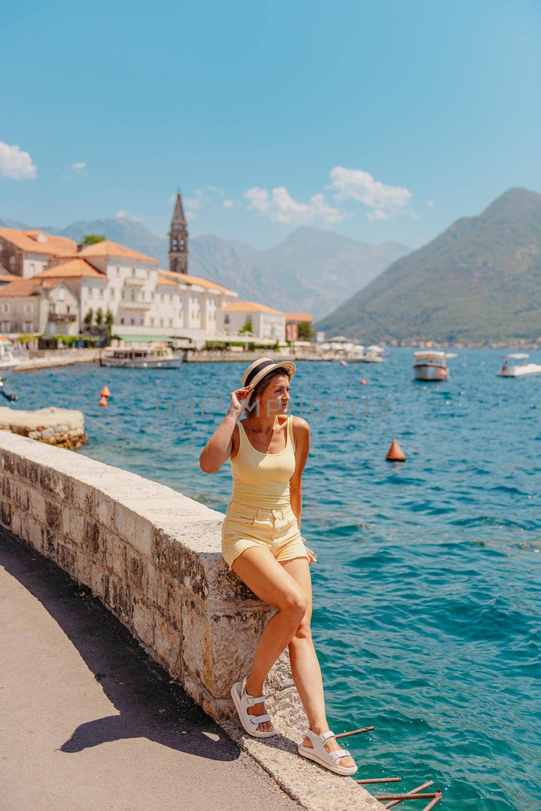 Summer photo shoot on the streets of Kotor, Montenegro. Beautiful girl in white dress and hat. smiling tourist girl with hat. Spectacular view of Montenegro with copy space. Ю fashion outdoor photo of beautiful sensual woman with blond hair in elegant dress and straw hat and bag, posing in Montenego's city Perast by Andrii_Ko