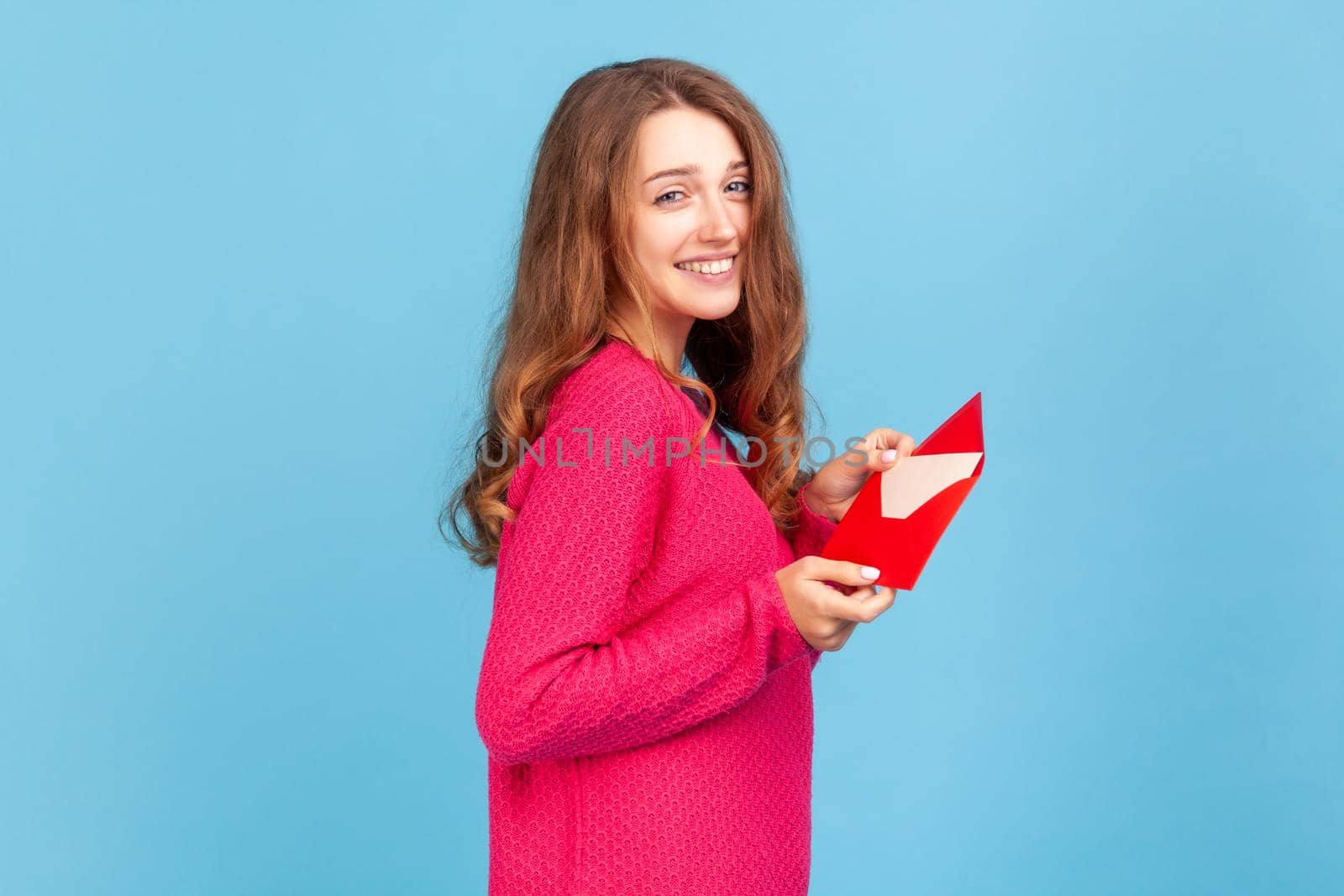 Side view portrait of woman wearing pink pullover, showing romantic letter in red envelope, holding love message and looking at camera with toothy smile. Indoor studio shot isolated on blue background