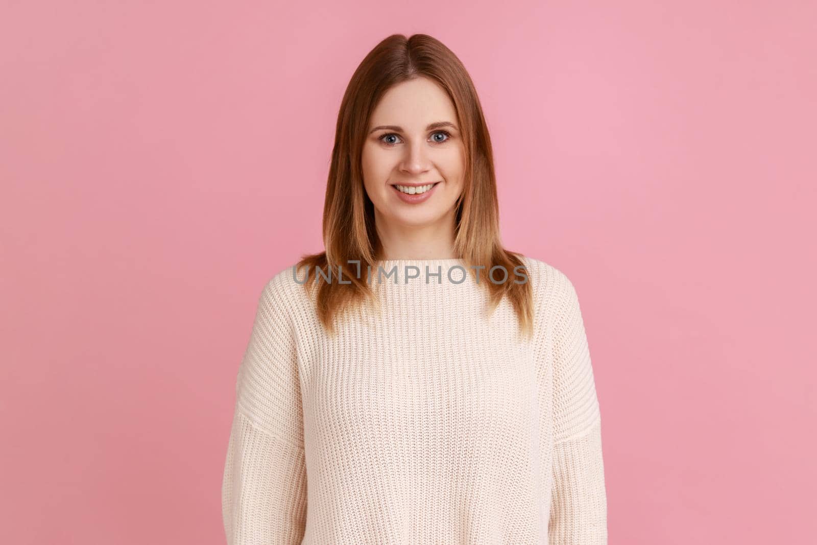 Portrait of positive successful blond woman standing with smile, looking happily at camera, expressing optimism, wearing white sweater. Indoor studio shot isolated on pink background.