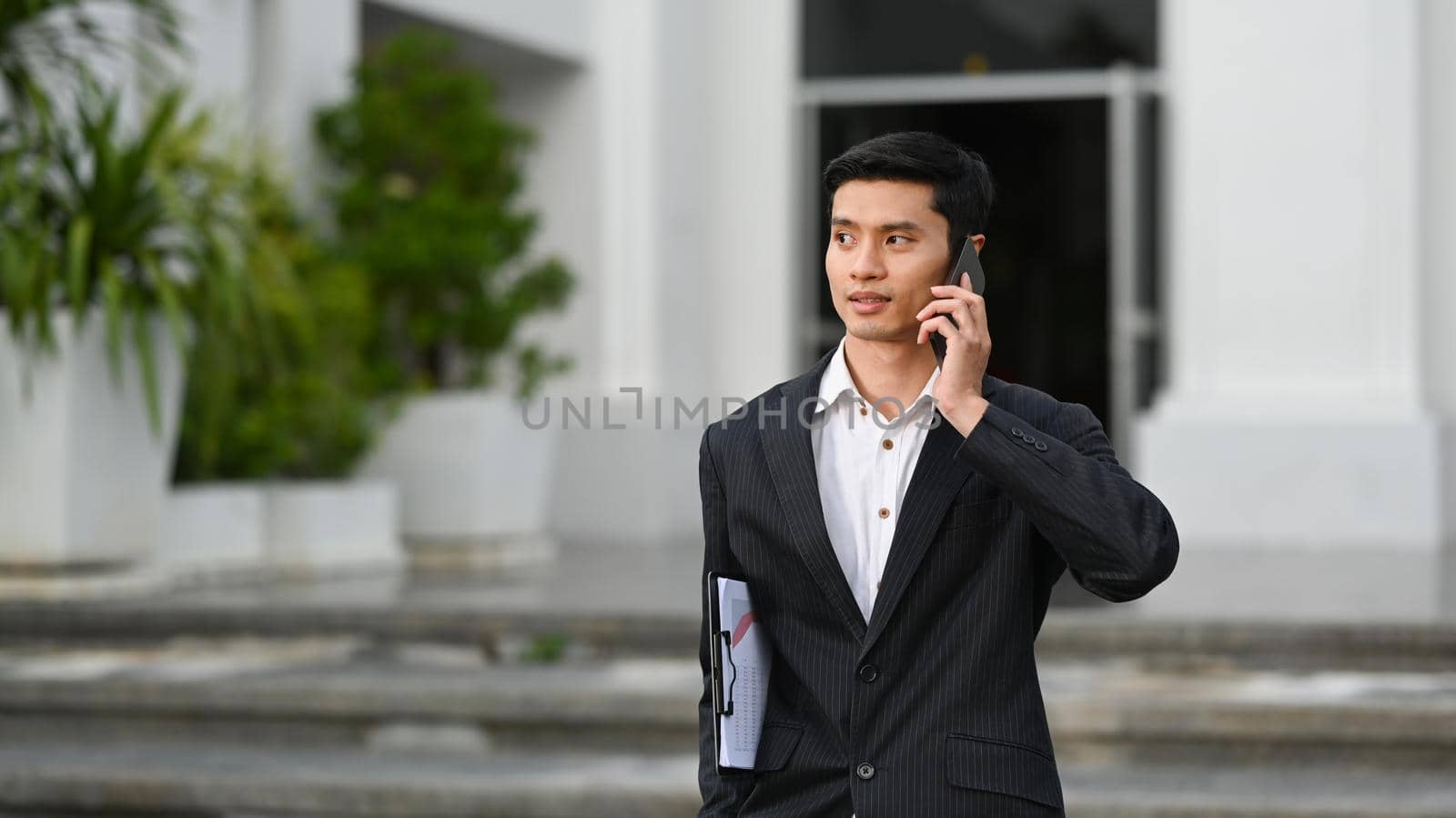 Young businessman in formal clothes standing in front of an office building and talking on mobile phone by prathanchorruangsak