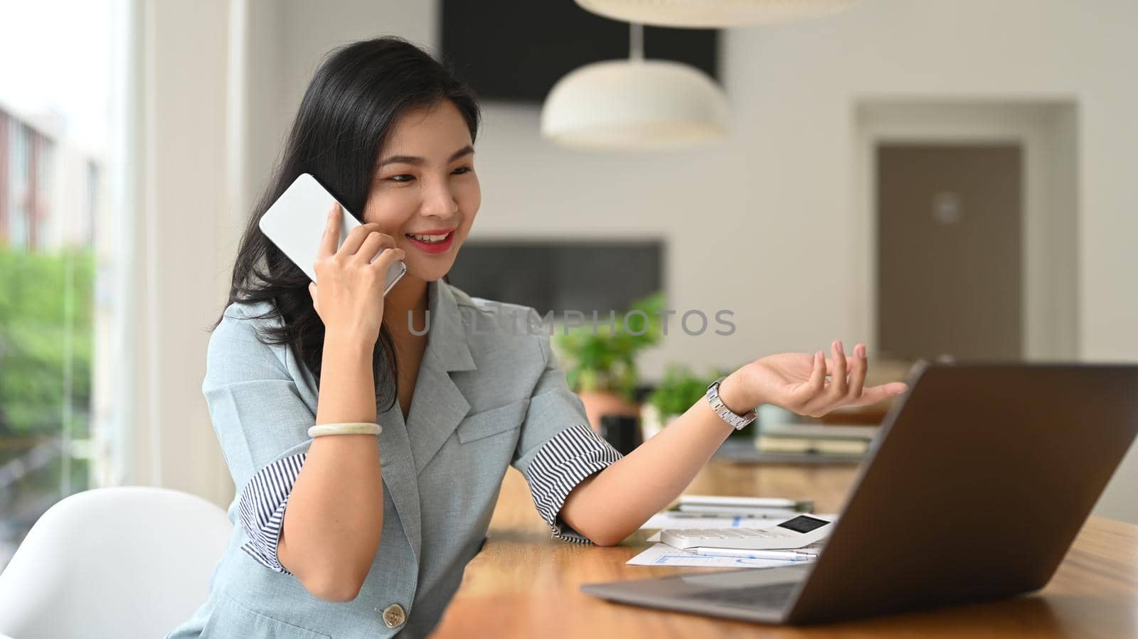 Asian female employee having pleasant phone conversation and using laptop at office desk by prathanchorruangsak
