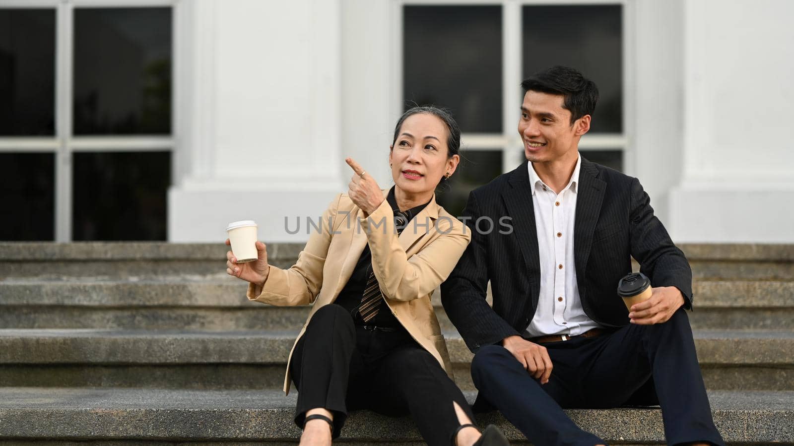 Mature businesswoman and young businessman talking each other, sitting on the stairs in front of business center by prathanchorruangsak