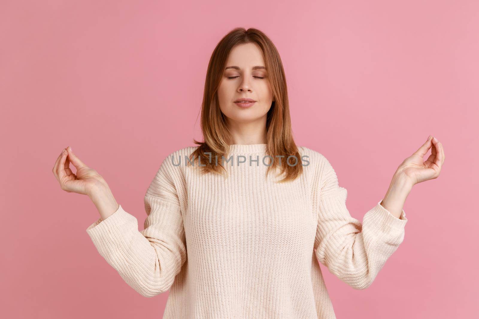 Portrait of blond woman closing eyes and holding hands up in mudra gesture, meditating, yoga exercise to reduce stress, wearing white sweater. Indoor studio shot isolated on pink background.