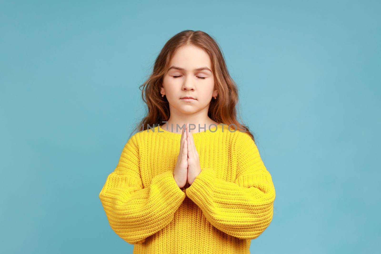 Portrait of little girl holding fingers in mudra gesture, feeling calm positive and relaxed, yoga practice, wearing yellow casual style sweater. Indoor studio shot isolated on blue background.