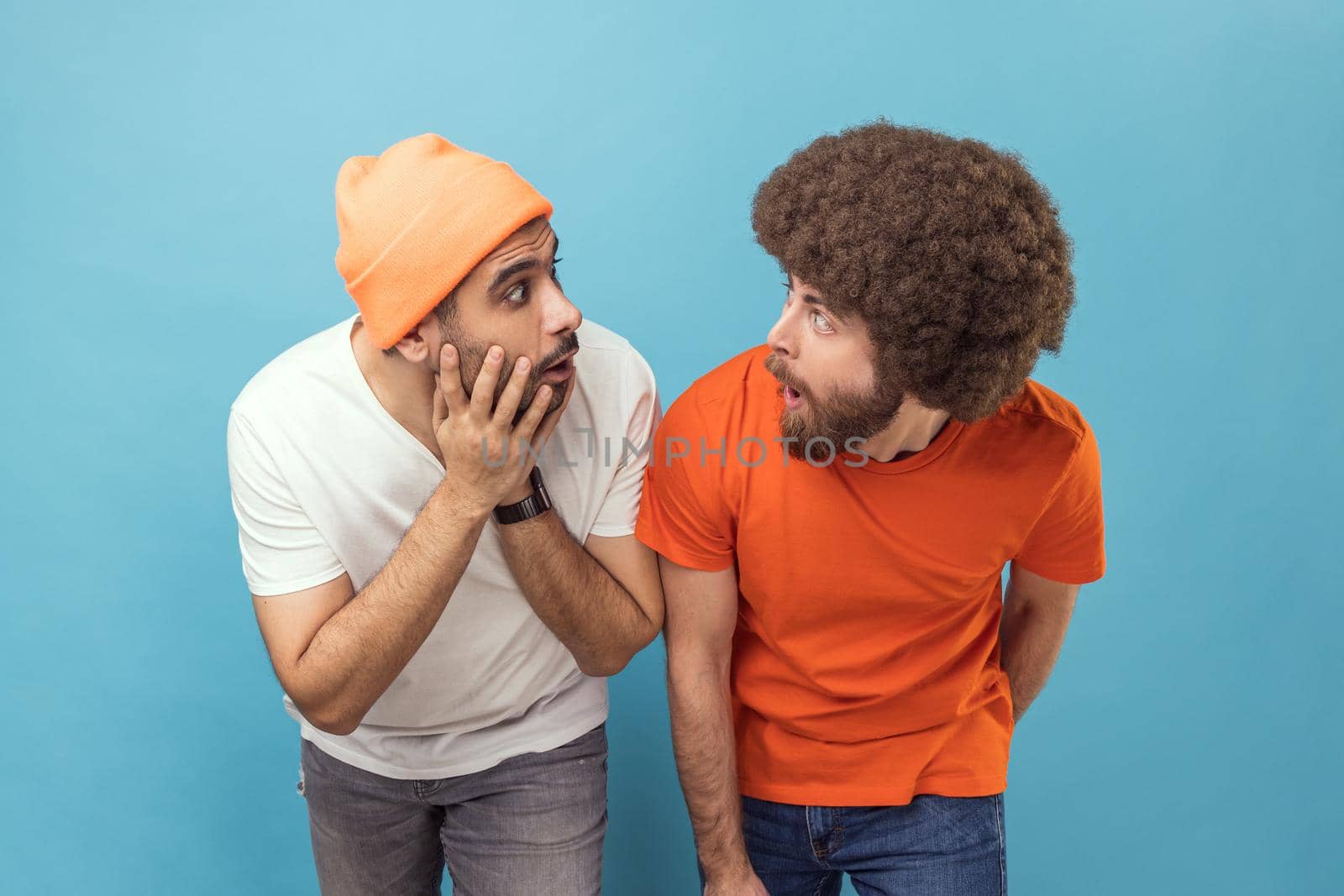 Portrait of two shocked amazed young adult hipster men standing looking at each other with big eyes and open mouth, being surprised. Indoor studio shot isolated on blue background.