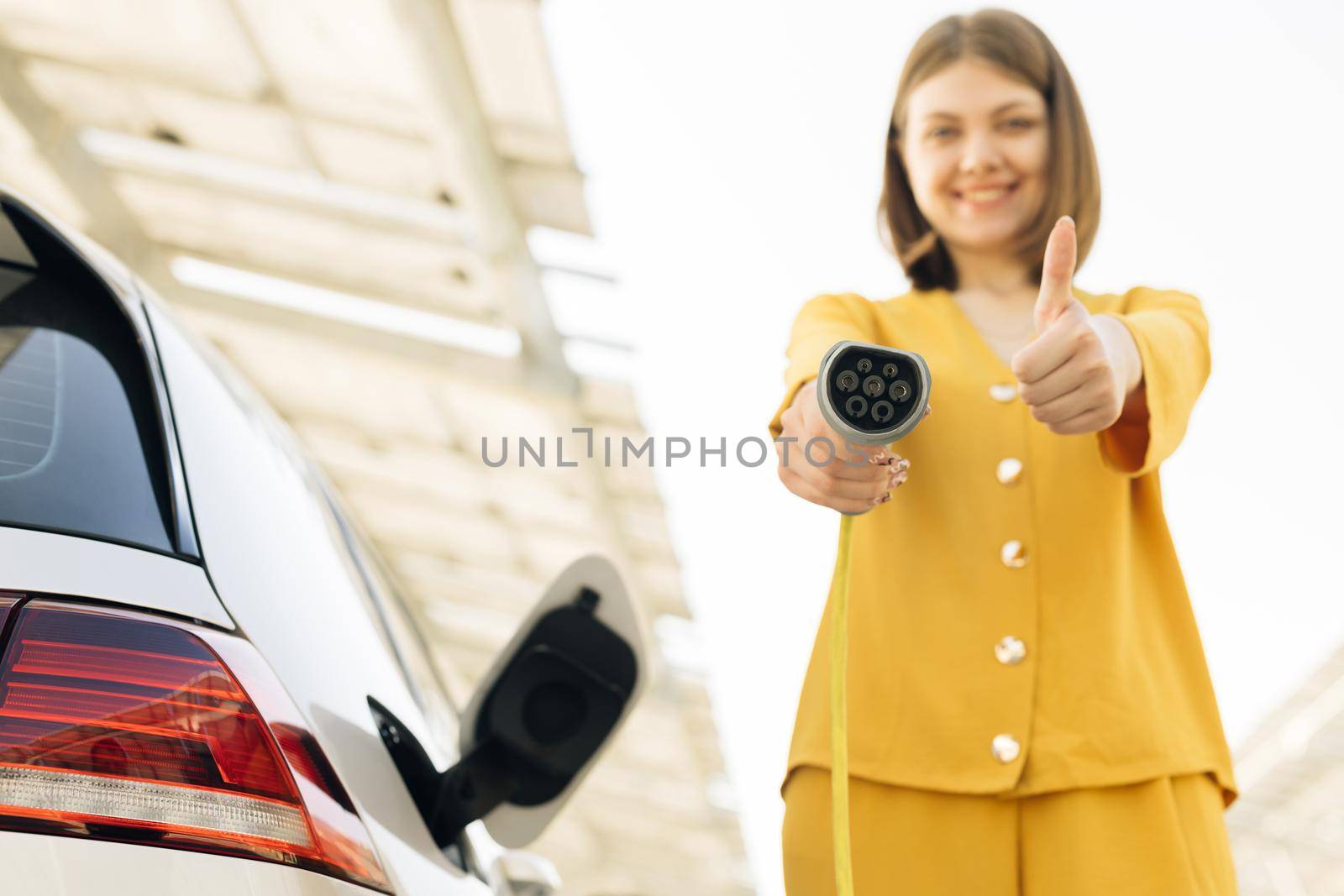 Female showing thumbs up holding power cable supply plugged at electric car charging station. Woman standing near electric charging station looking at camera and showing thumb up.