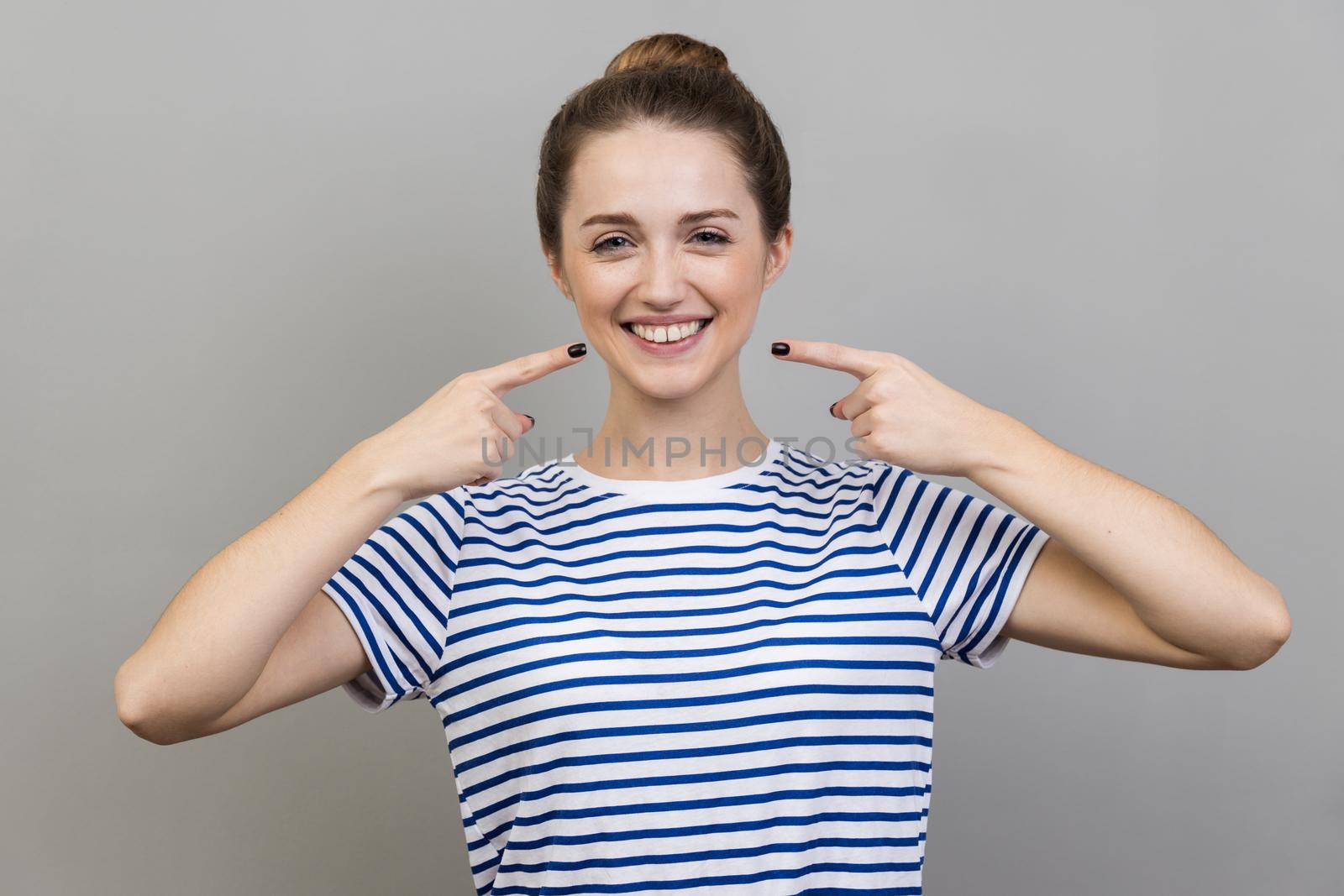 Portrait of pretty cheerful woman wearing striped T-shirt with bun hairstyle points index fingers at smile shows white teeth, looking at camera. Indoor studio shot isolated on gray background.