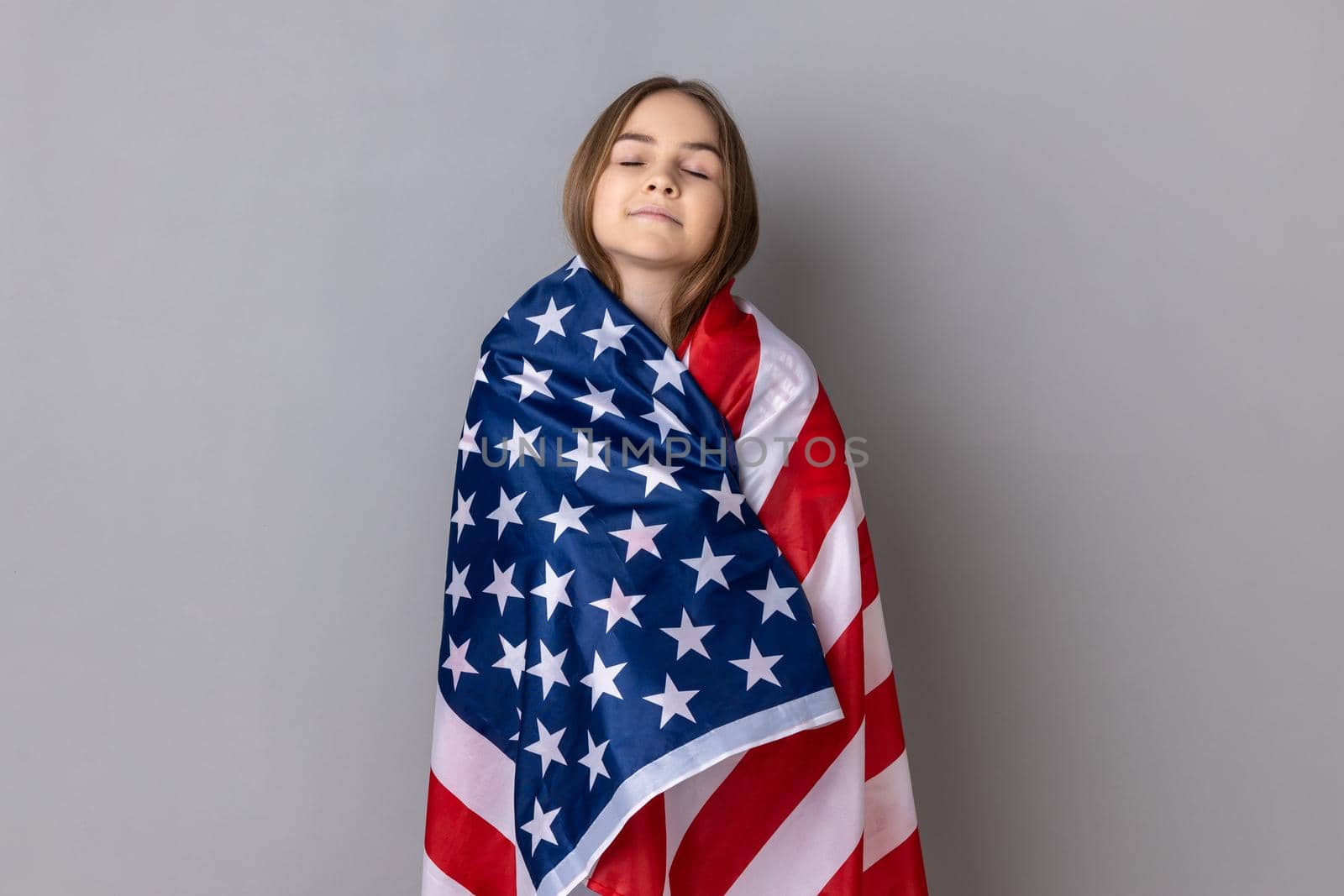 Portrait of dark haired patriotic little girl wearing striped T-shirt standing wrapped in american flag, keeping eyes closed, relocating to America. Indoor studio shot isolated on gray background.