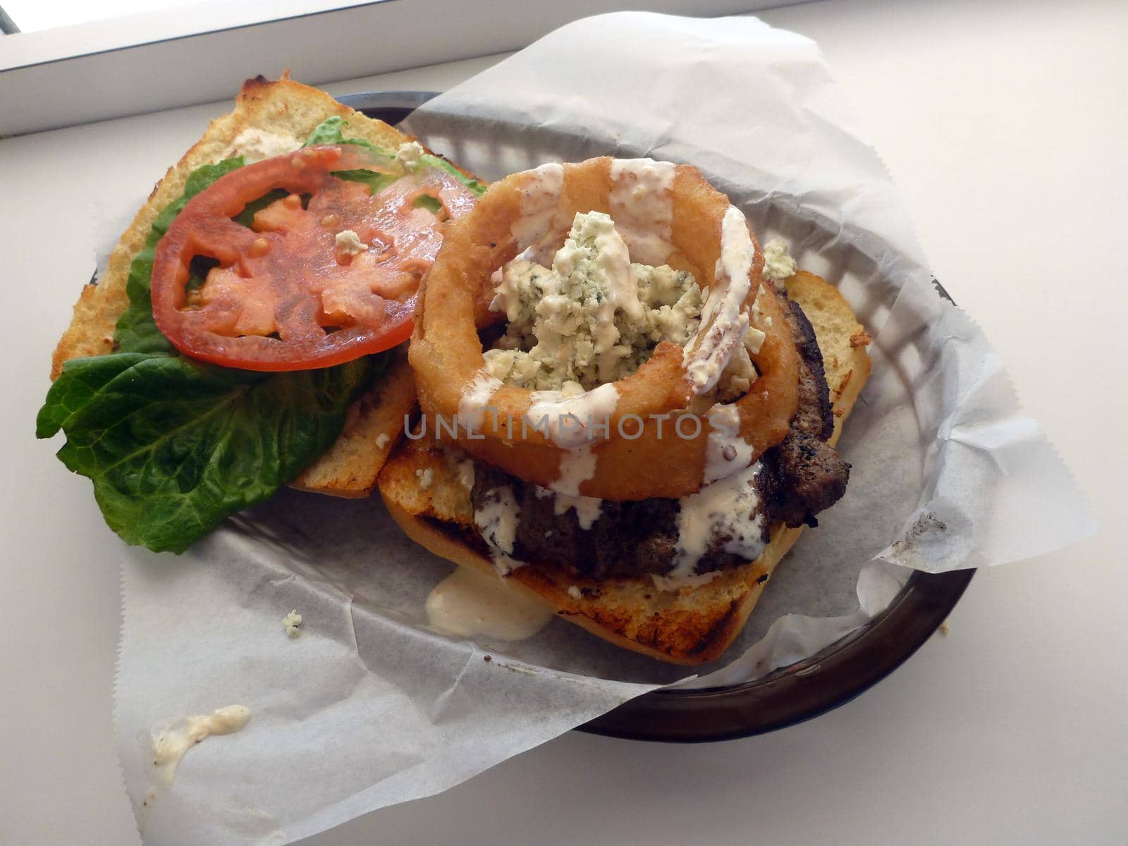 "Blue Holes" Burger Onion Rings, Blue Cheese crumbles, Ciabatta, Aioli, Lettuce and tomato in a plastic basket on a white table.