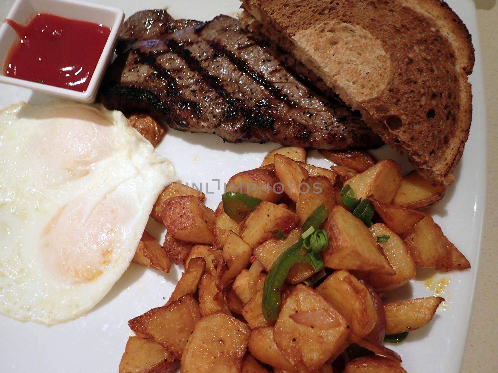 Steak with two eggs over easy, two slices of rye bread, potatos and cup of ketchup on square plate with steak knife on table.