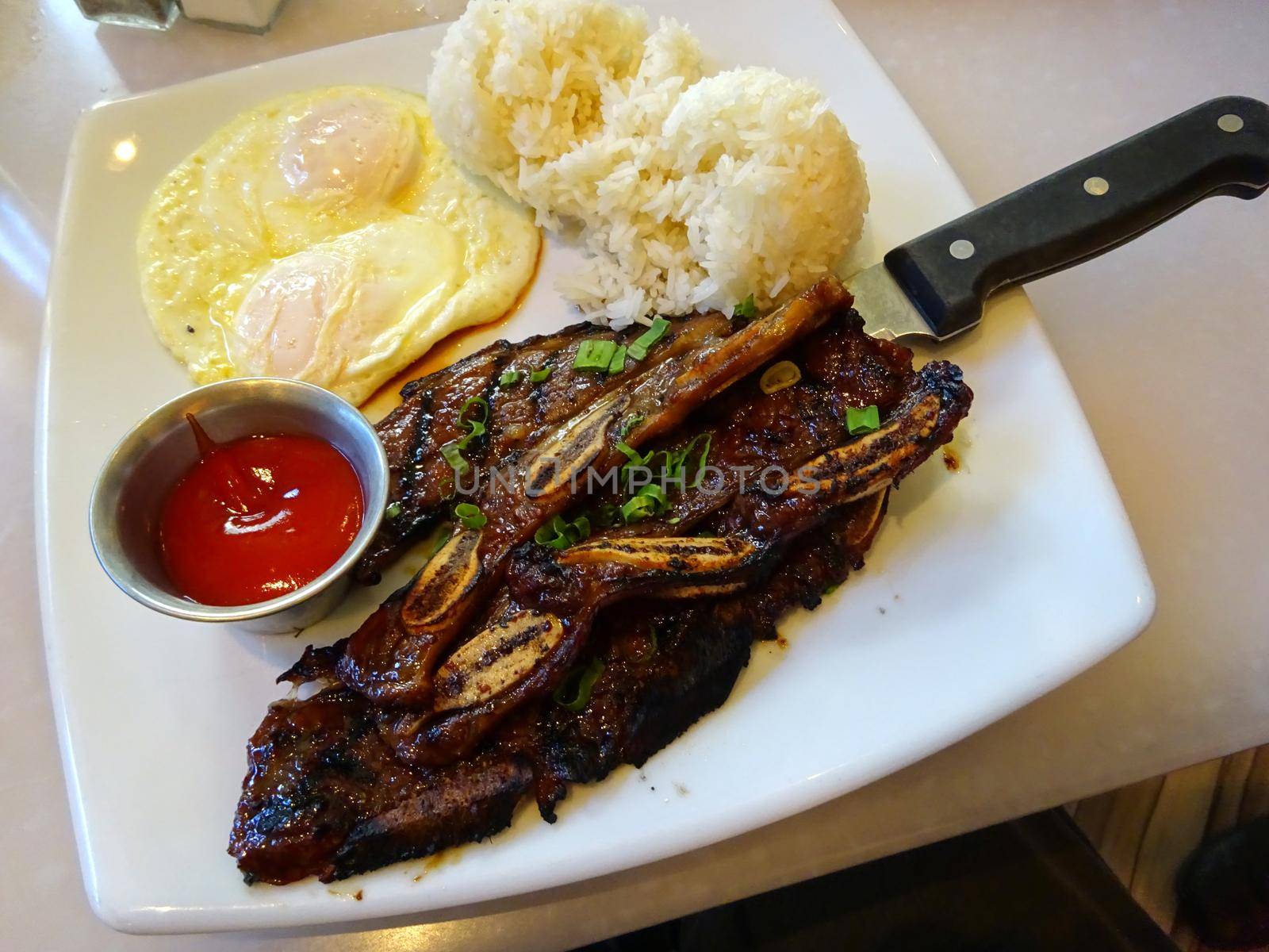 Kalbi steak ribs with two eggs over easy, two scoops of rice and cup of ketchup on square plate with steak knife on table.