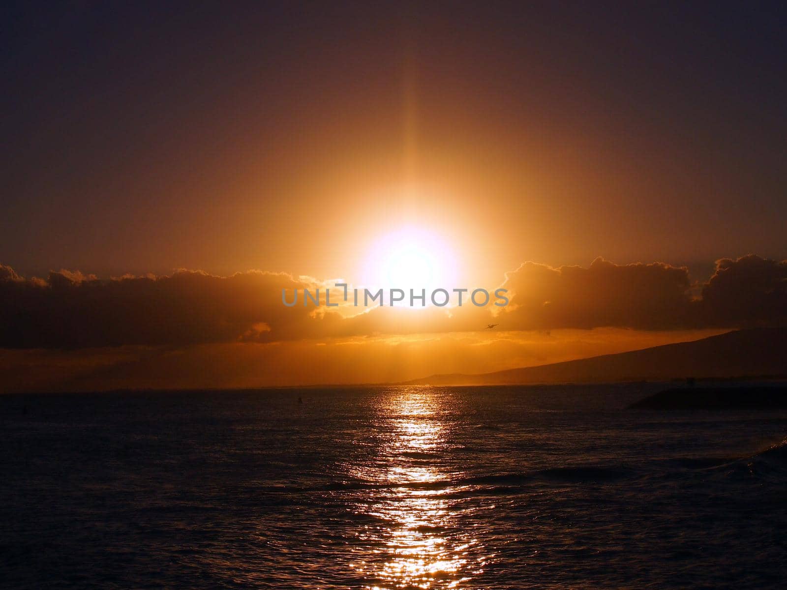 Dramatic Sunset over the clouds and reflecting on the Pacific ocean on the water with airplane flying off the coast of Oahu, Hawaii with Waianae Mountain range visible.