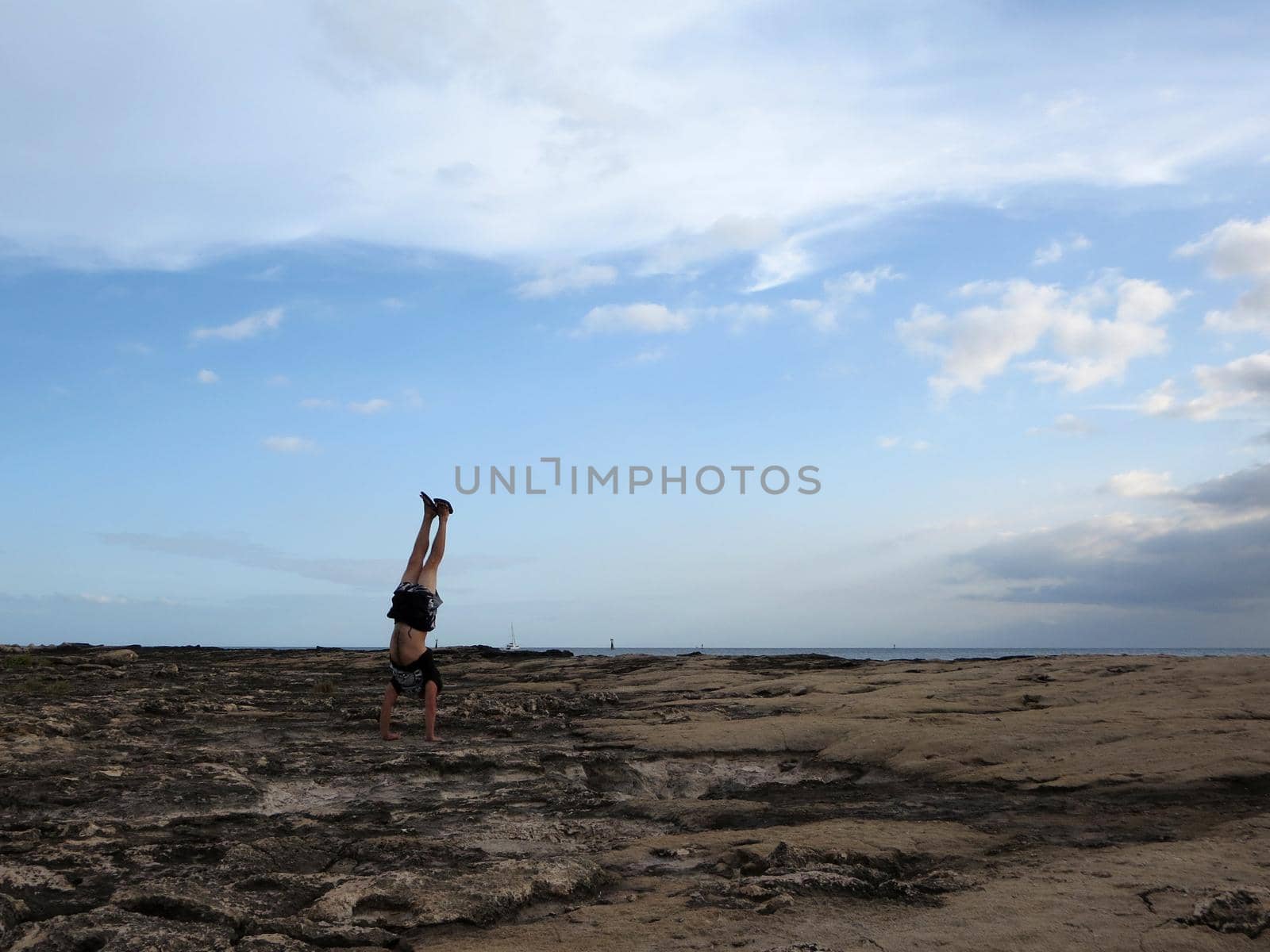 Man wearing a t-shirt, shorts, and slippers Handstands on shore rocks of Ko Olina on Oahu, Hawaii with dramatic clouds, sky, and pacific ocean in the distance. 