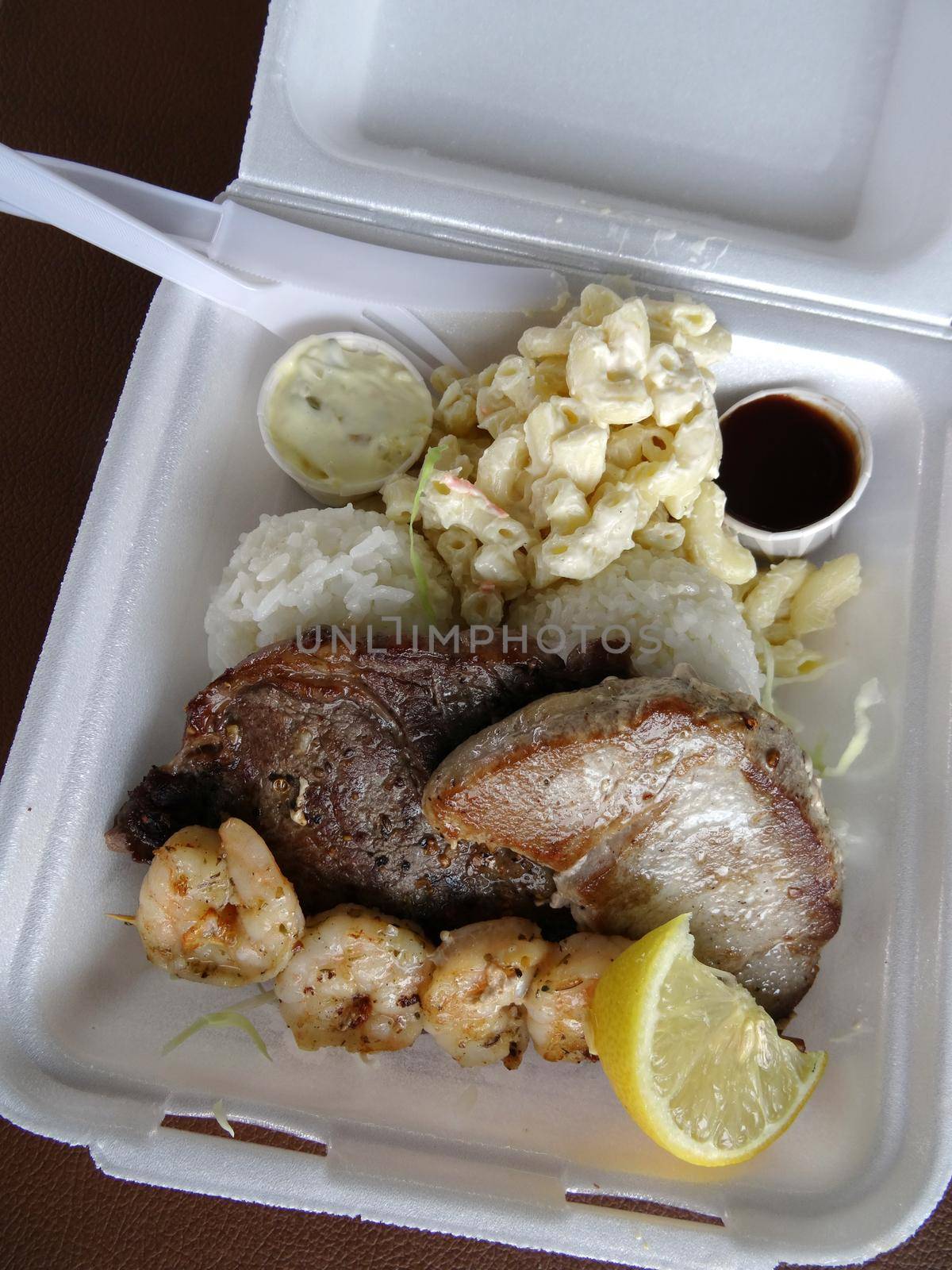 Surf & Turf Includes: Ahi, Steak, Shrimp, rice, and macaroni  salad with small cups of sauce, lemon in a styrofoam tray with plastic knife and fork.