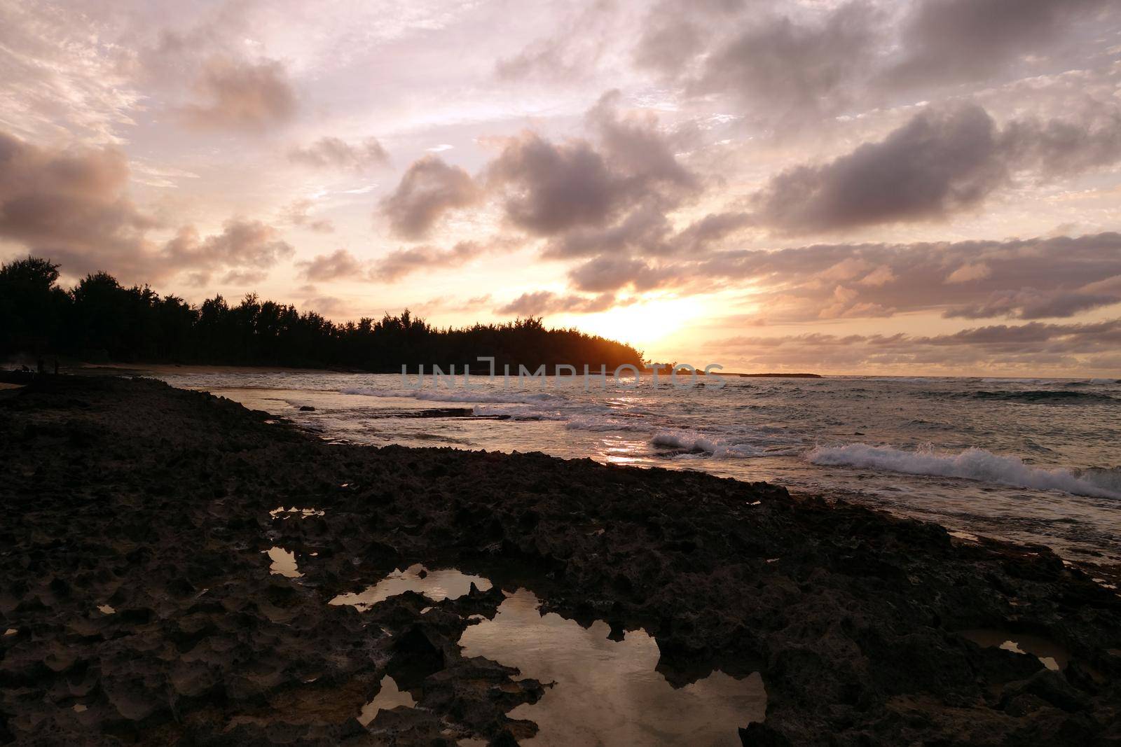 Sunset through the clouds and reflecting on the waves as they break on rocky shore with tide pools at Kawela Bay on the North Shore of Oahu. 