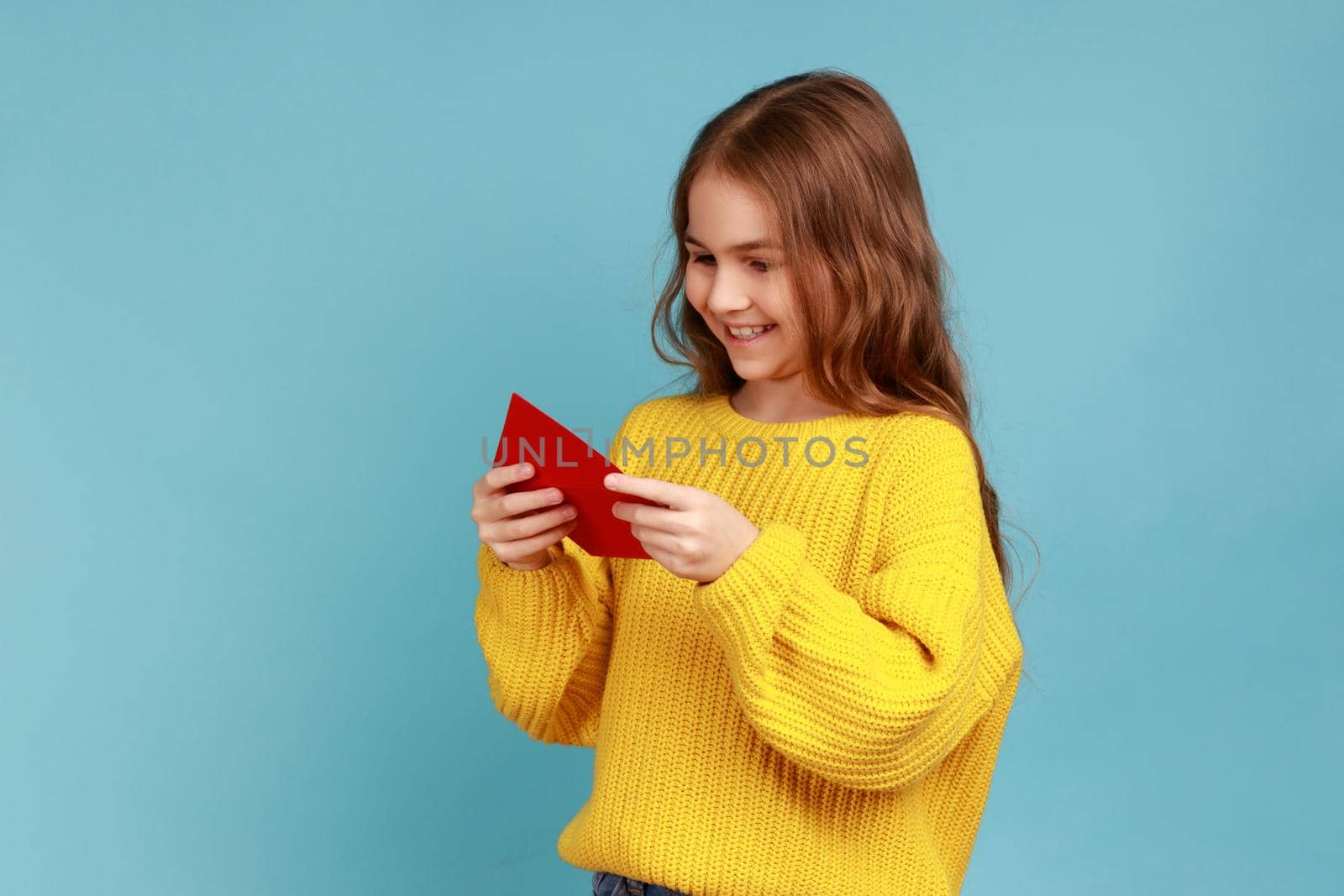 Portrait of little girl holds red envelope in hands, reads letter from classmate with charming smile, wearing yellow casual style sweater. Indoor studio shot isolated on blue background.