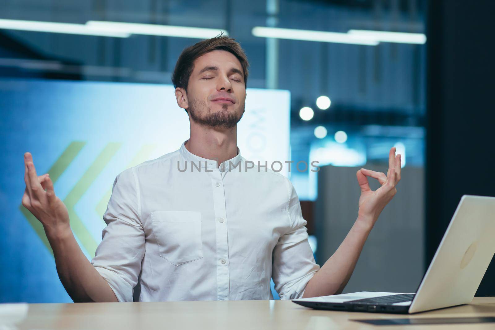 Young handsome man freelancer, businessman, manager in the office at the table resting, closed his eyes, meditates, shows his hands yoga poses.